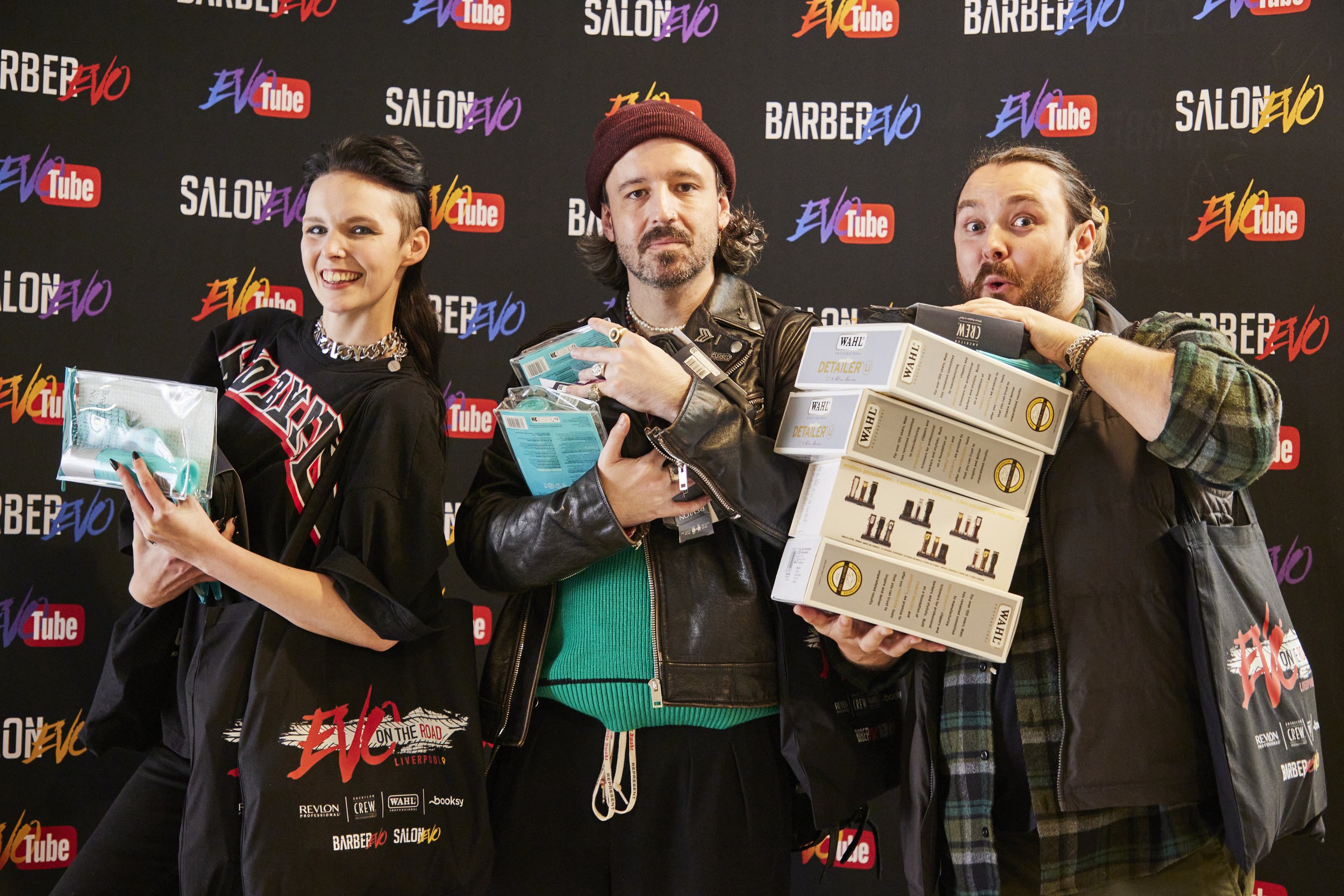 Three people holding products and posing in front of a black backdrop with logos for SalonEvo, YouTube, and Barbershop. The person on the left is a woman with dark hair holding hair products and wearing a black shirt with branding. The person in the