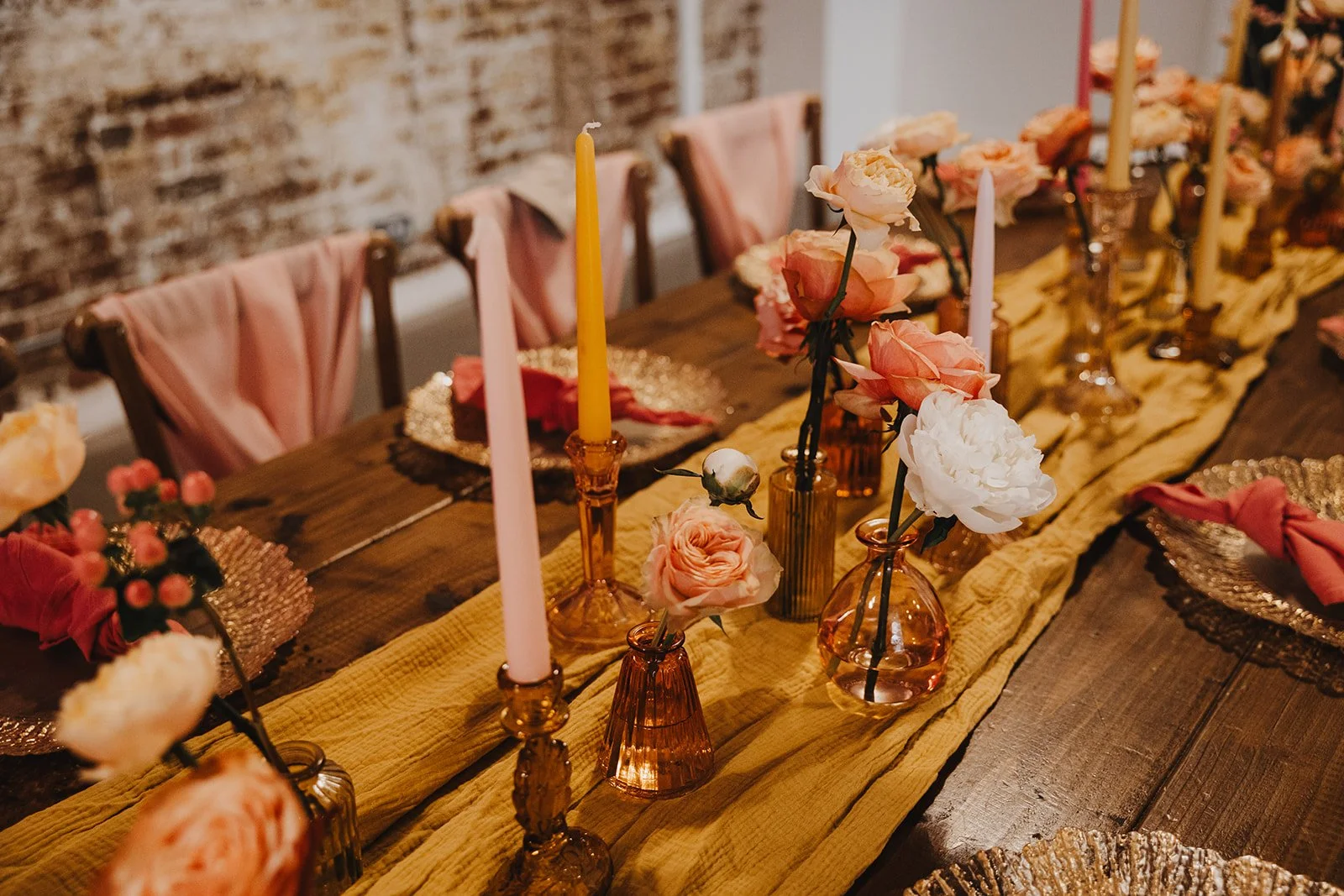 Decorated dining table with pink and white roses in small vases, pink and yellow candles, and pink napkins on transparent plates, with chairs draped in pink cloth.