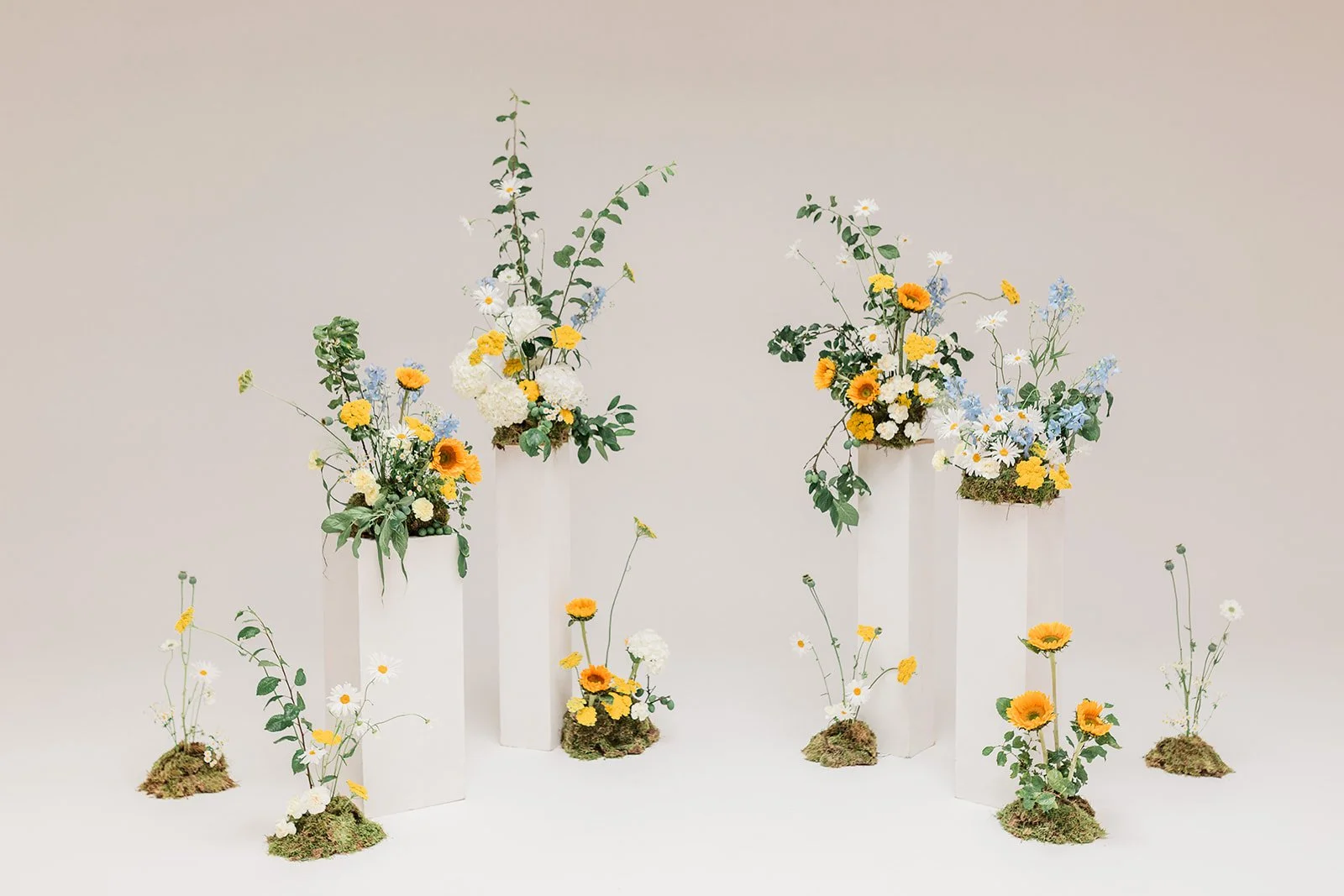 Arrangement of colorful floral bouquets with white, yellow, and blue flowers in white vases and small moss-covered arrangements on a white background.