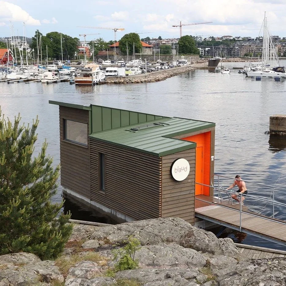 En flytende badstue med grønt tak som flyter på vannet ved en brygge i Kristiansand. En person går opp en badestige. Det er en marina med mange båter og havnebygninger i bakgrunnen.