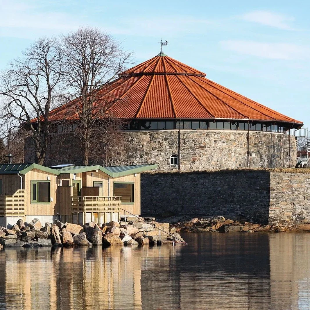 Christiansholm festning i Kristiansand plassert ved vannkanten. Badstue med moderne arkitektur. Furukledning med store vinduer og en terrasse, omgitt av steiner og vann.