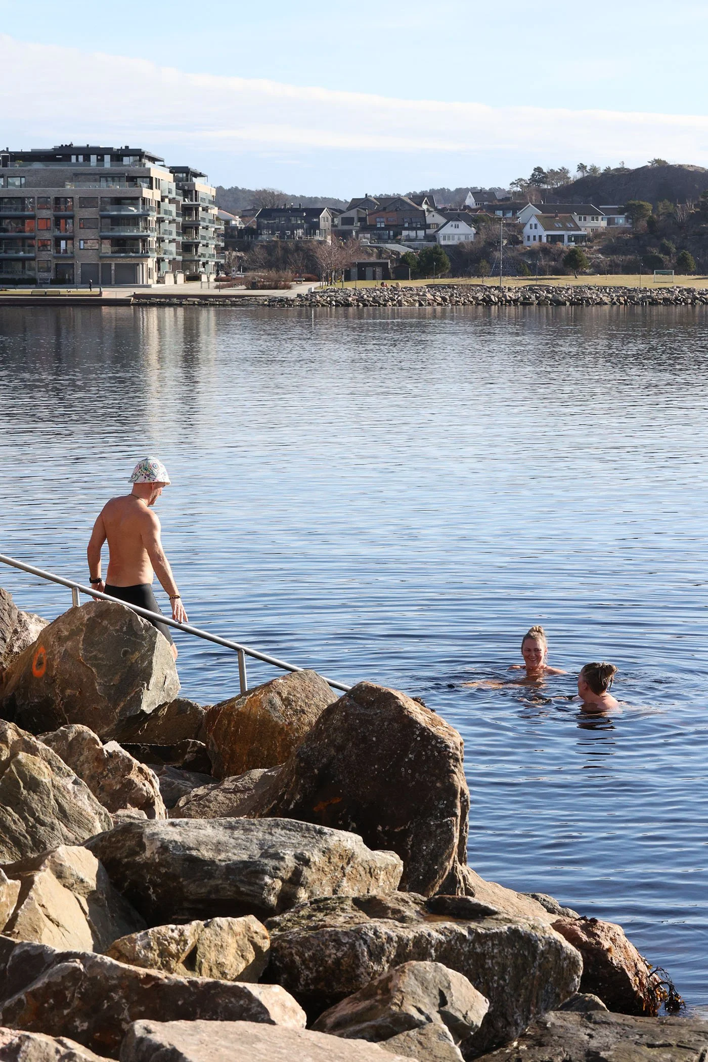 En person med badedrakt og badetøy går ut av vannet, mens to personer svømmer og ler i vannet ved en steinbrygge ved en innrammet bygrunn med boligblokker og hytter på åsene.