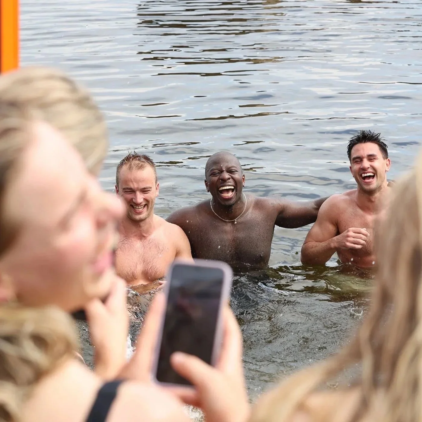 Tre menn som ler og smiler mens de står i vannet ved en badstue.