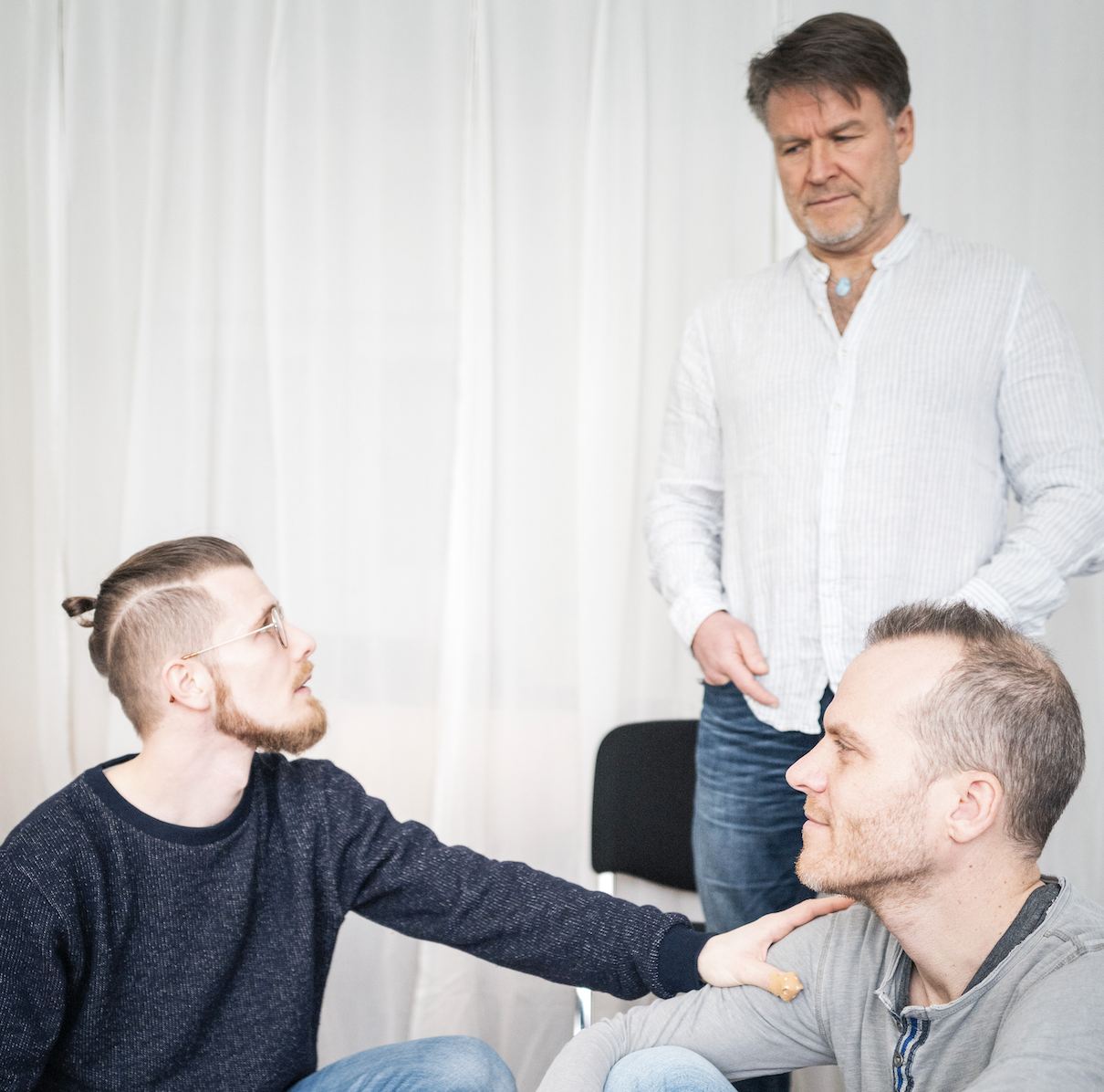 Zwei Männer sitzen sich gegenüber, einer legt eine Hand auf die Schulter des anderen, während ein dritter Mann im Hintergrund zuschaut, in einem Raum mit weißen Vorhängen.