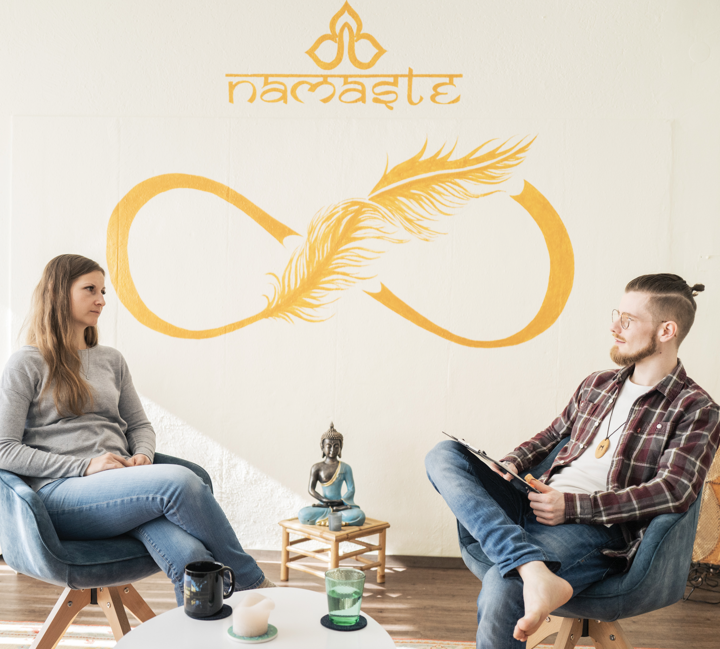 Zwei Menschen sitzen sich in einem Beratungsraum gegenüber, im Hintergrund eine Wand mit einem gelben Symbol eines Unendlichkeitssymbols, darüber das Wort 'namaste' und eine goldene Federschleife. Zwischen ihnen steht ein kleines Beistelltischchen mit einer Statue, Kerze und einer Keramikschale. Auf dem Tisch vor ihnen befinden sich eine schwarze Tasse und eine grüne Glasflasche. Der Raum wirkt entspannt und einladend.