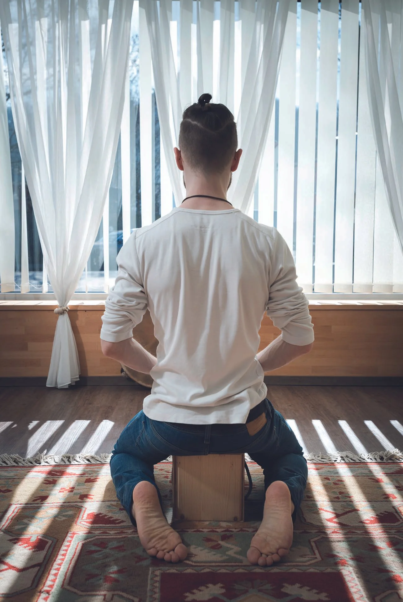Raphael Komanns sitzt auf einem kleinen Hocker vor einem Fenster mit weißen Vorhängen, die Licht in den Raum lassen.
