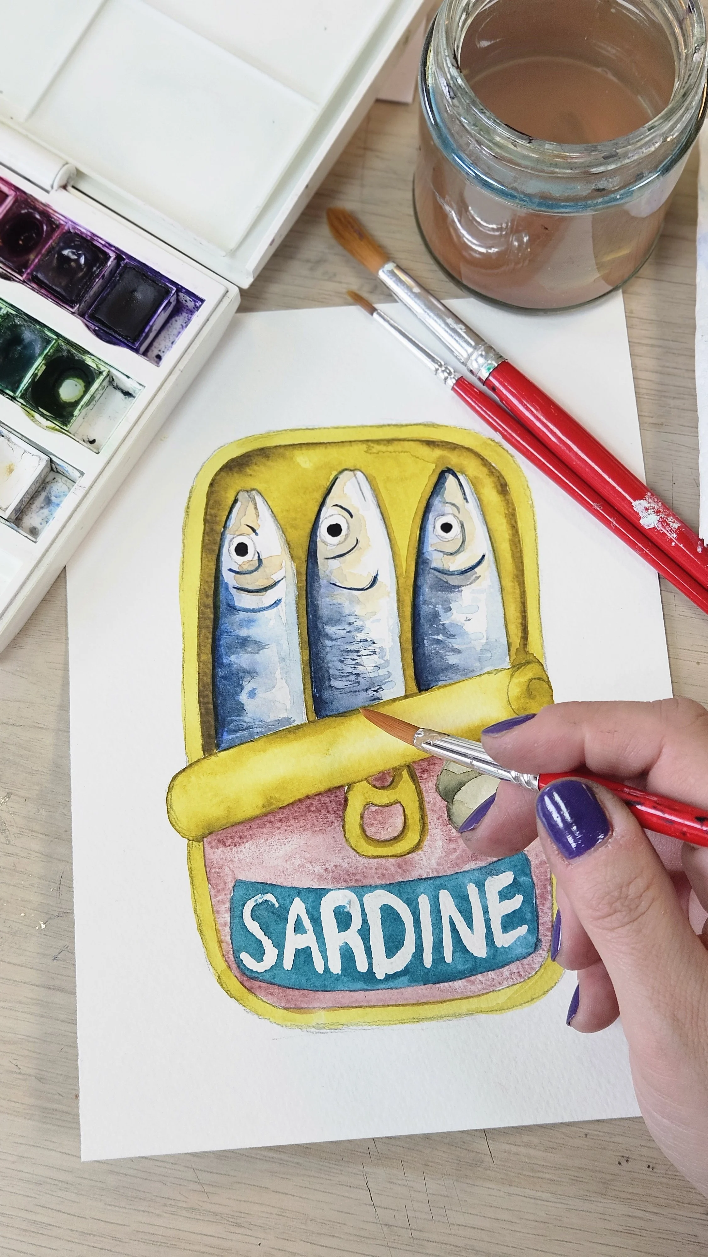 A person painting a watercolor illustration of a sardine can labeled 'SARDINE'. The illustration features three sardines with blue and silver bodies inside a yellow can with a pull-tab top. Two paintbrushes and a jar of water are on the table, along with watercolor paints.