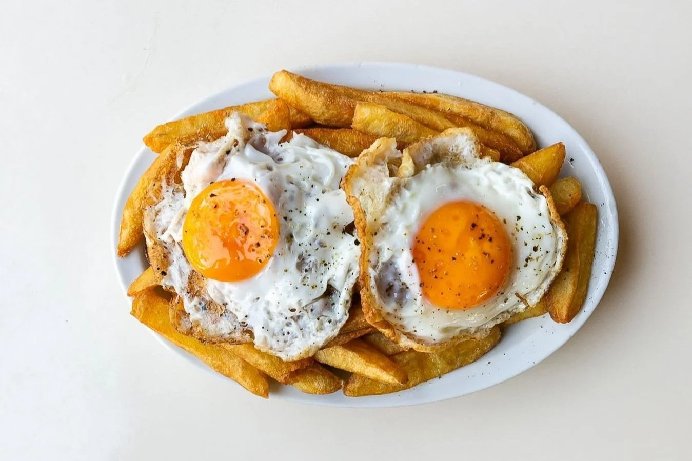 Fried eggs with sunny side up on top of French fries served on a white plate.