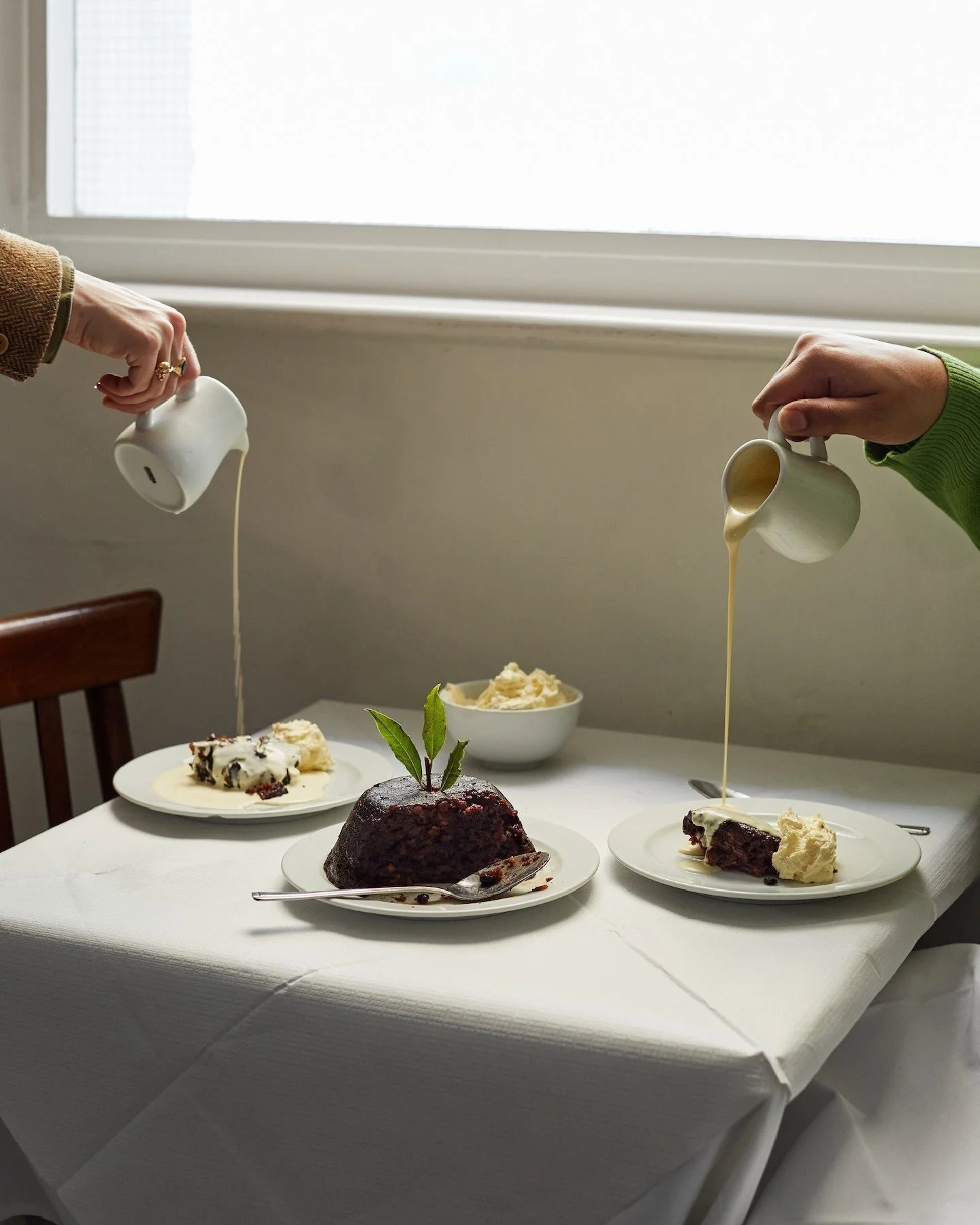 Two people pouring cream over slices of cake and a whole cake with a green garnish on a table.