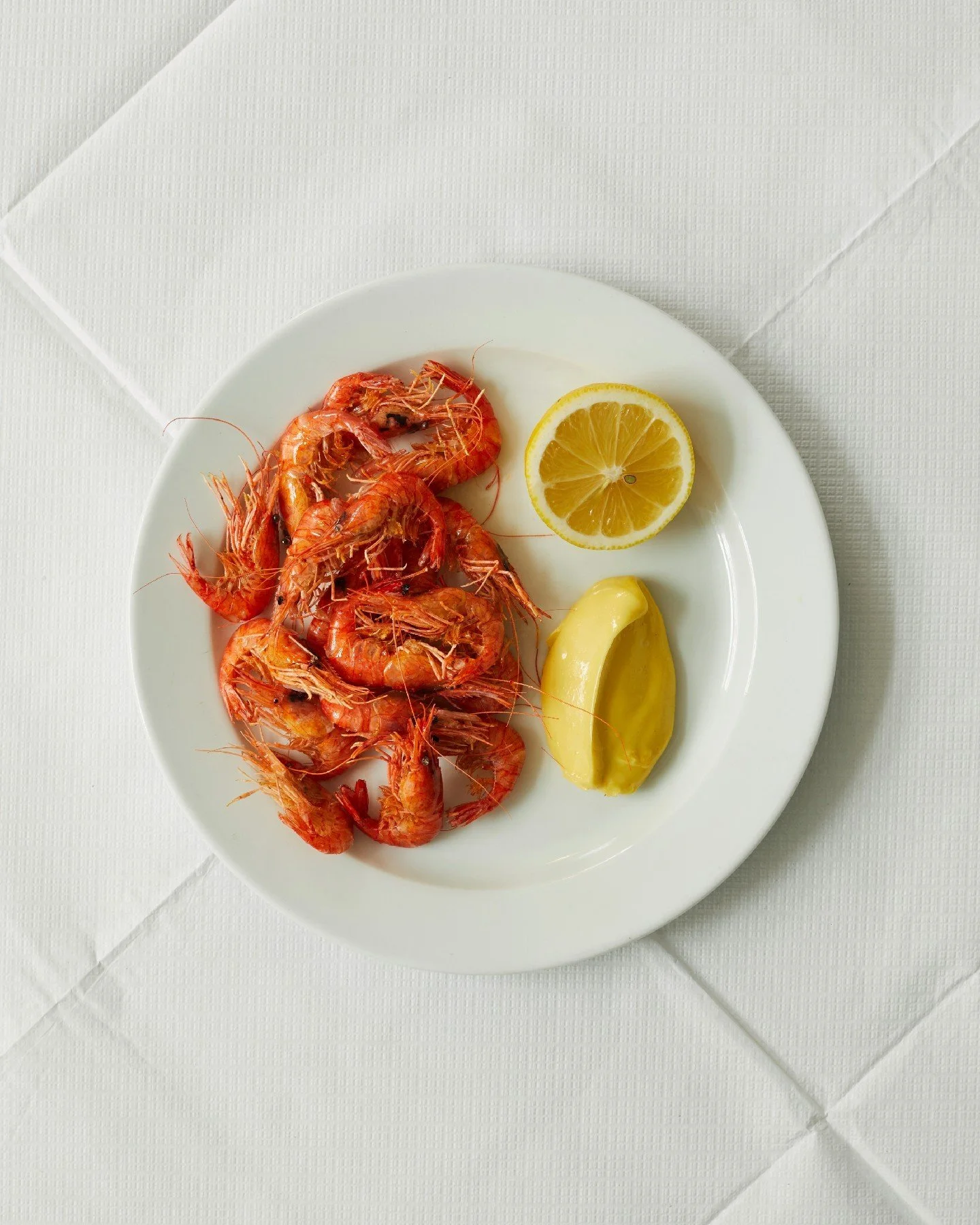 Plate of cooked shrimp with lemon slice and a dollop of butter on a white plate, set on a white tablecloth.