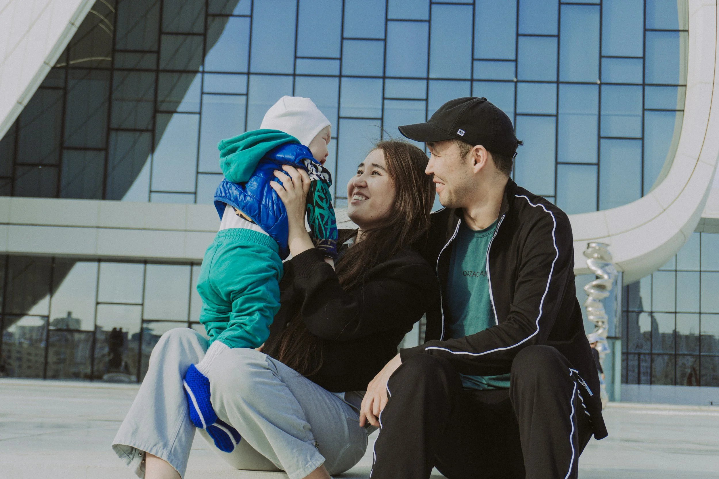 A family of three, including a little boy, sitting outside a modern glass building. The mother and father are smiling at each other, with the mother holding the boy's face, creating a warm, happy scene.