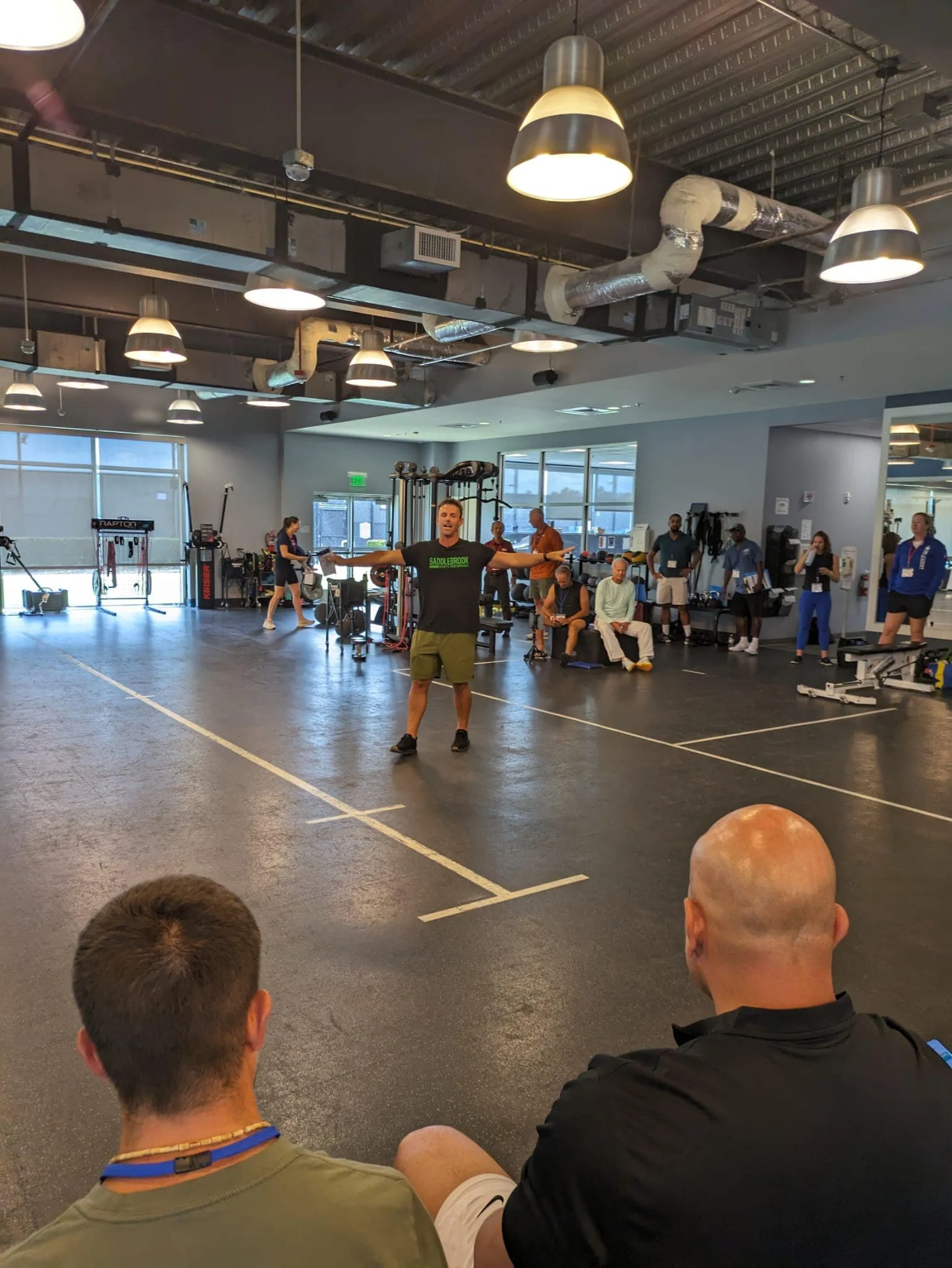 A man in a black t-shirt and shorts standing inside a gym with his arms outstretched, facing seated individuals, with other people and gym equipment in the background.