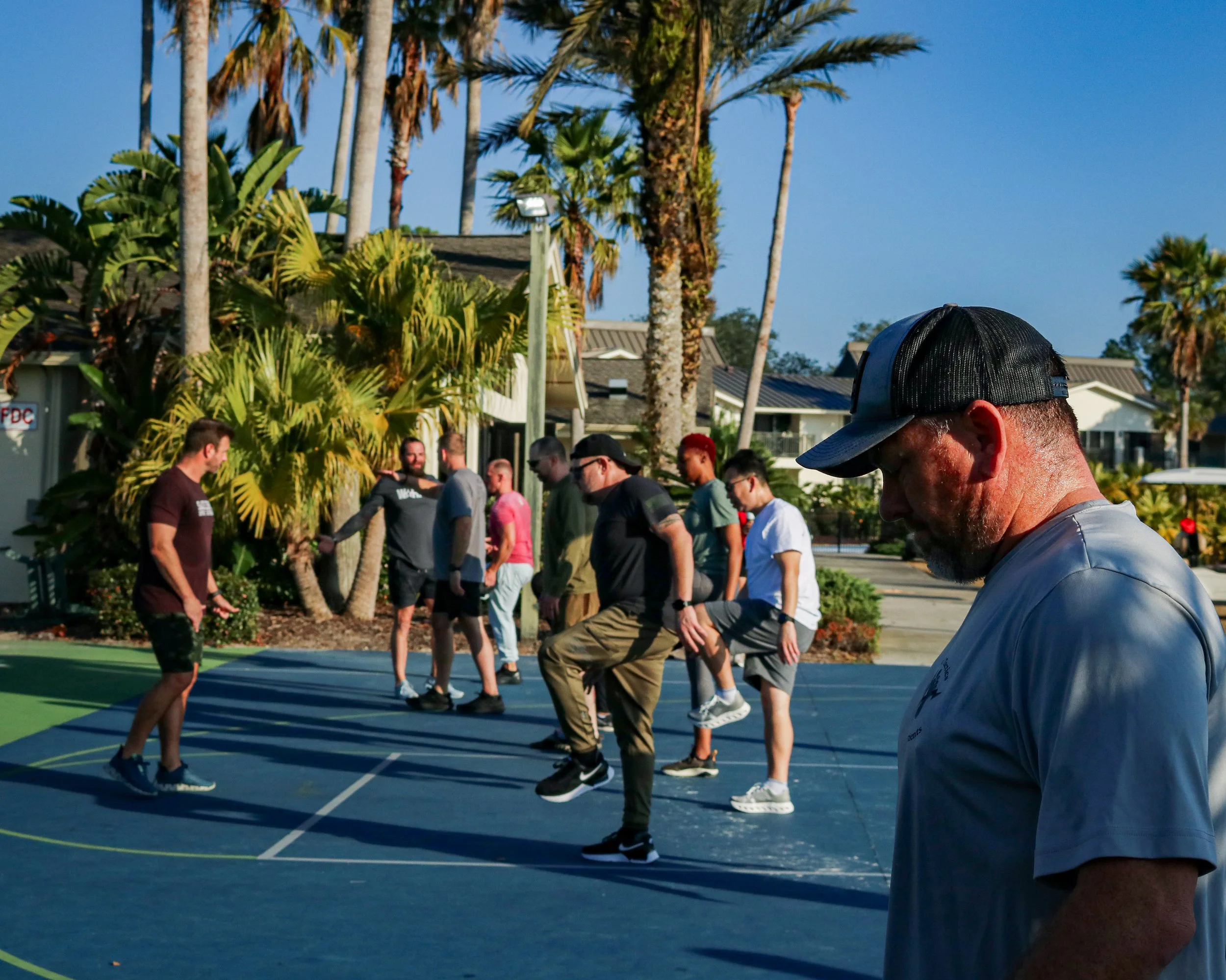 A group of people standing on a court, some stepping or balancing on lines, with palm trees and buildings in the background on a sunny day.