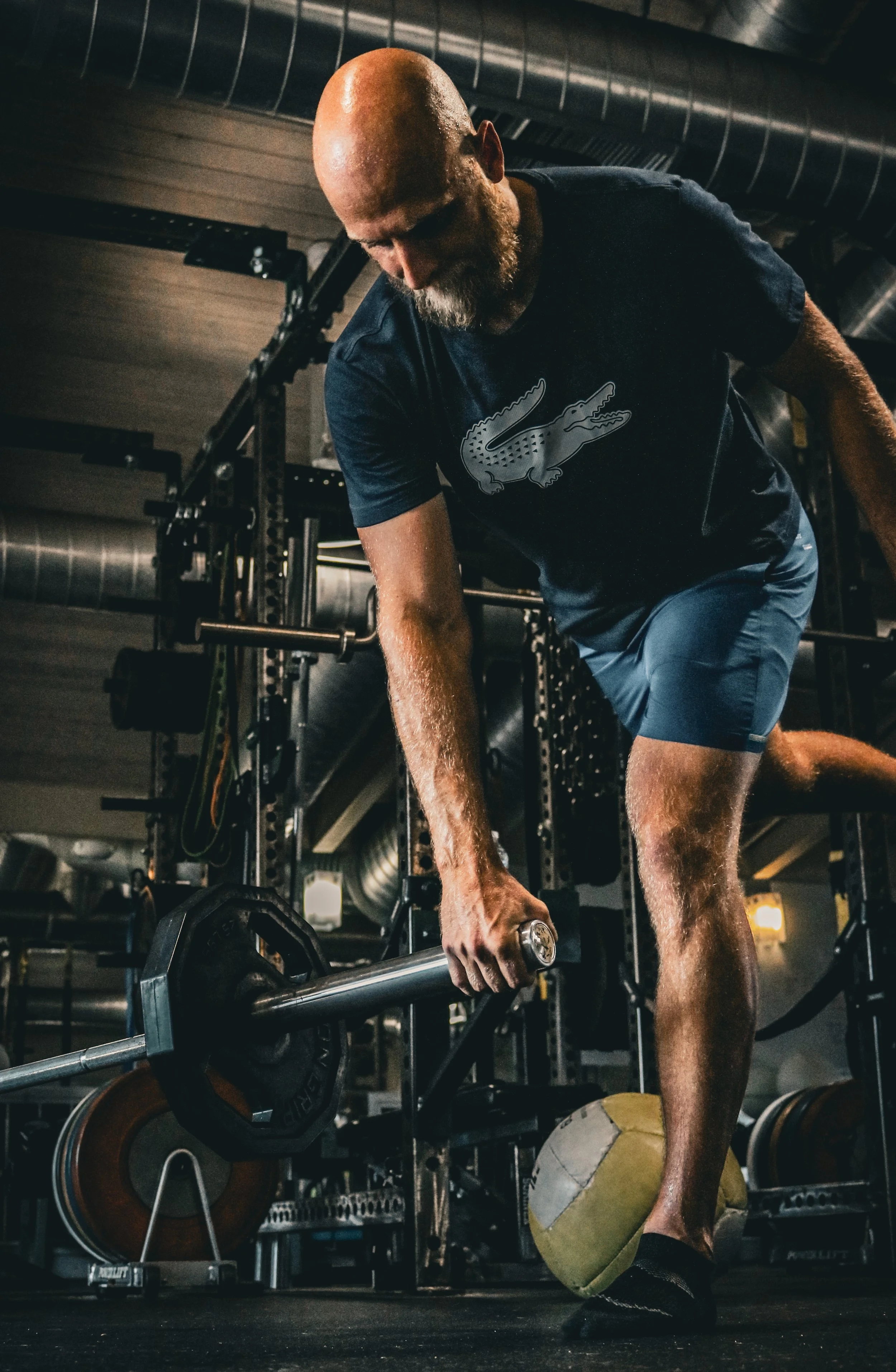 A man in a gym lifting a barbell with weights. He is wearing a dark t-shirt with a crocodile logo and blue shorts, standing on one leg with a medicine ball under his foot.