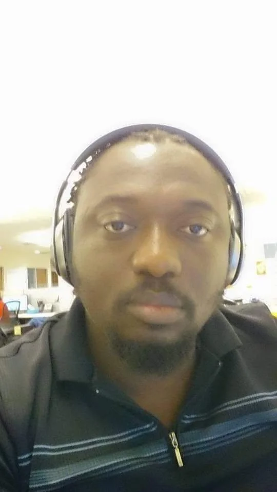 Close-up of a man with headphones, wearing a black zip-up jacket with white stripes, in an indoor setting with tables and chairs in the background.