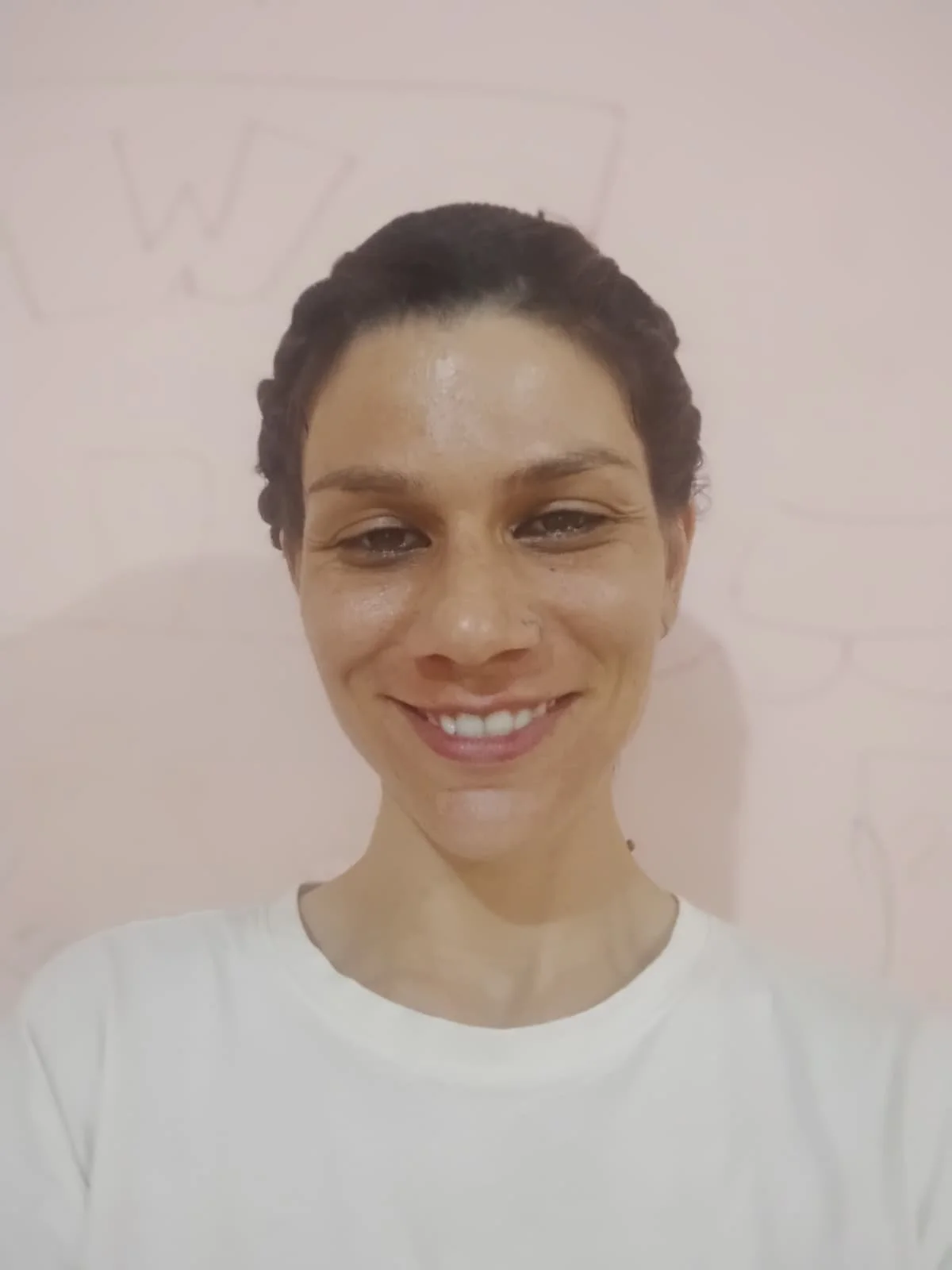 A woman smiling, with dark hair styled in braids, wearing a white top, standing in front of a light-colored wall with faint writing and a drawing in the background.