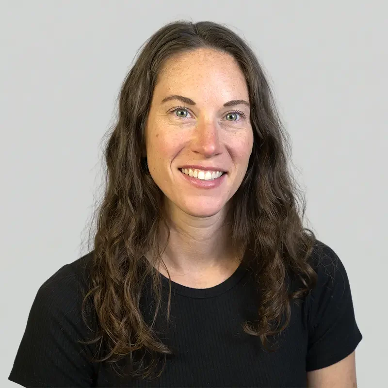 A woman with long, curly brown hair and blue eyes smiling at the camera, wearing a black t-shirt against a plain light-colored background.