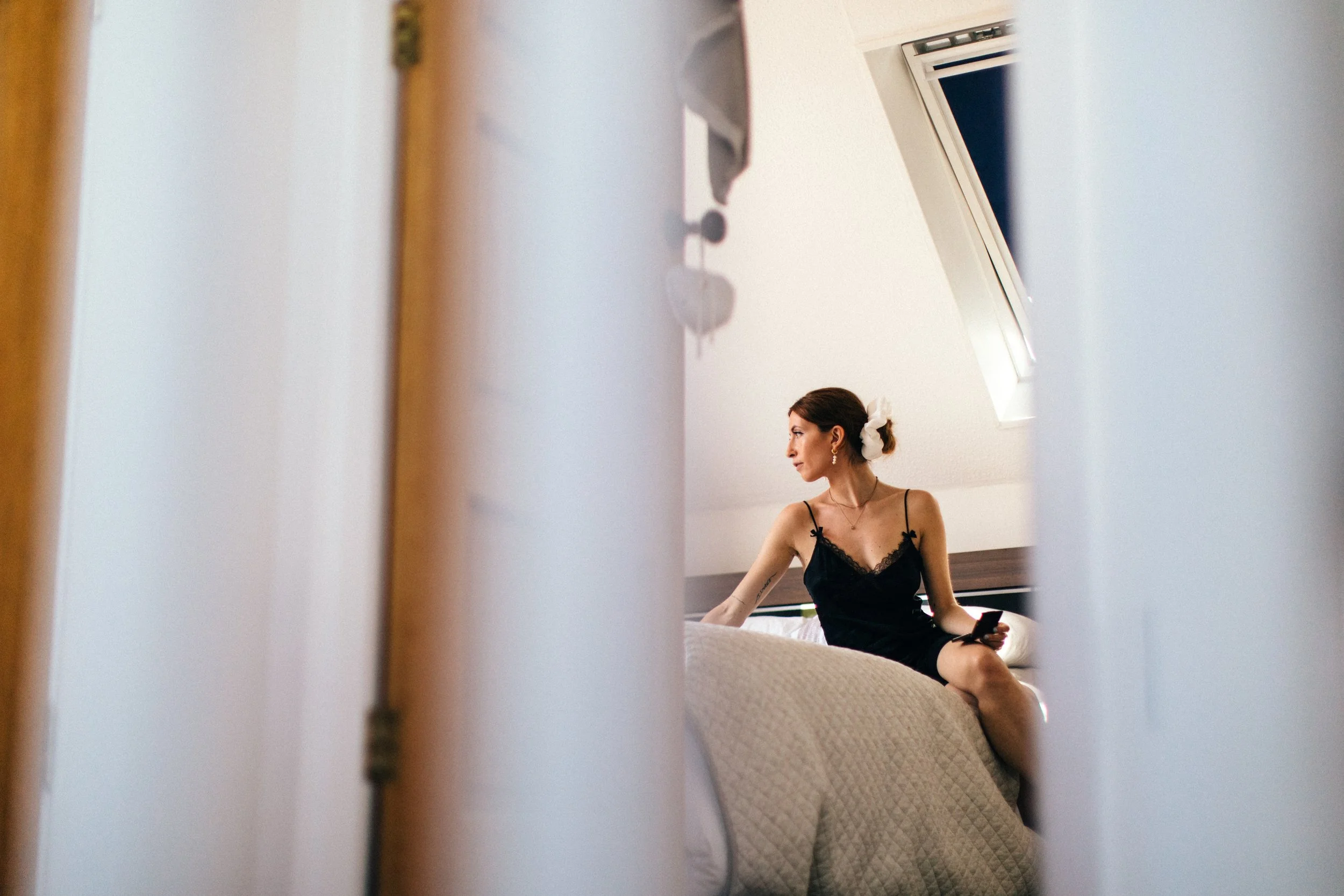 A bride sitting on a bed in a bedroom, looking to her left, wearing a black spaghetti strap dress, with her hair tied back and a white hair accessory, getting ready for her wedding day.