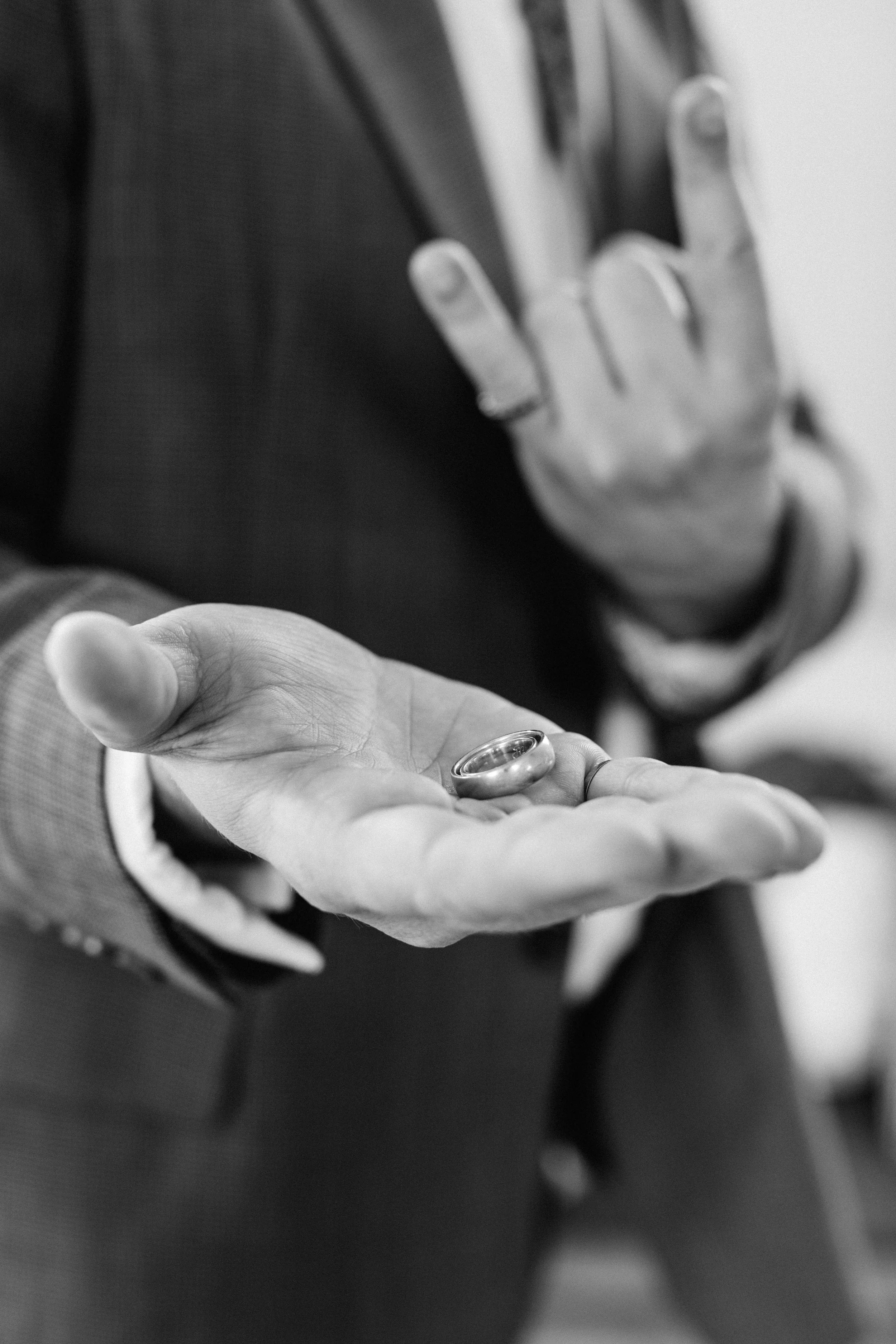 a Best Man in a suit holds a ring in their open palm, with another hand making a gesture in the background.
