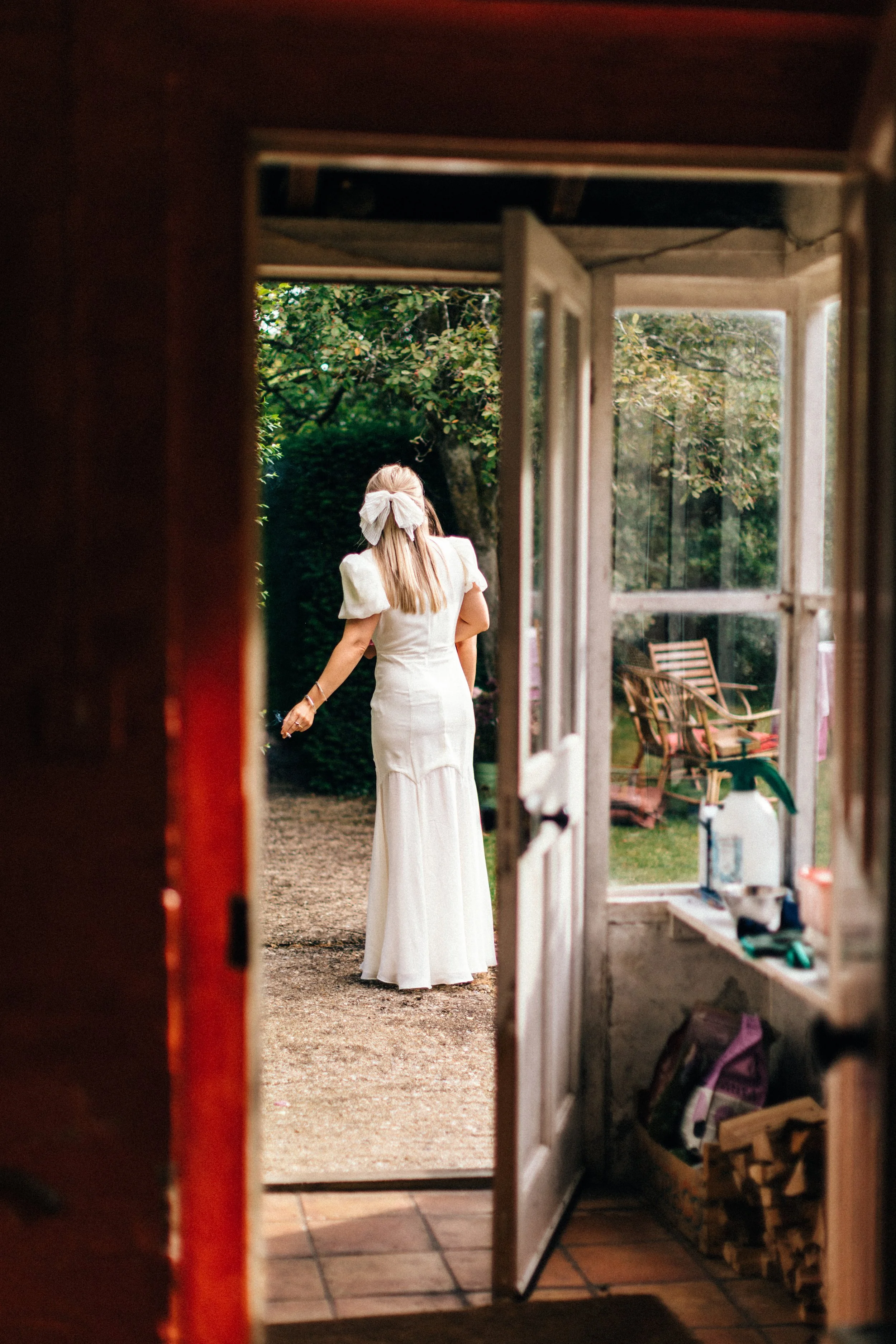 A woman in a white dress with a large bow in her hair, standing outside in a garden, viewed from inside a house through an open door.