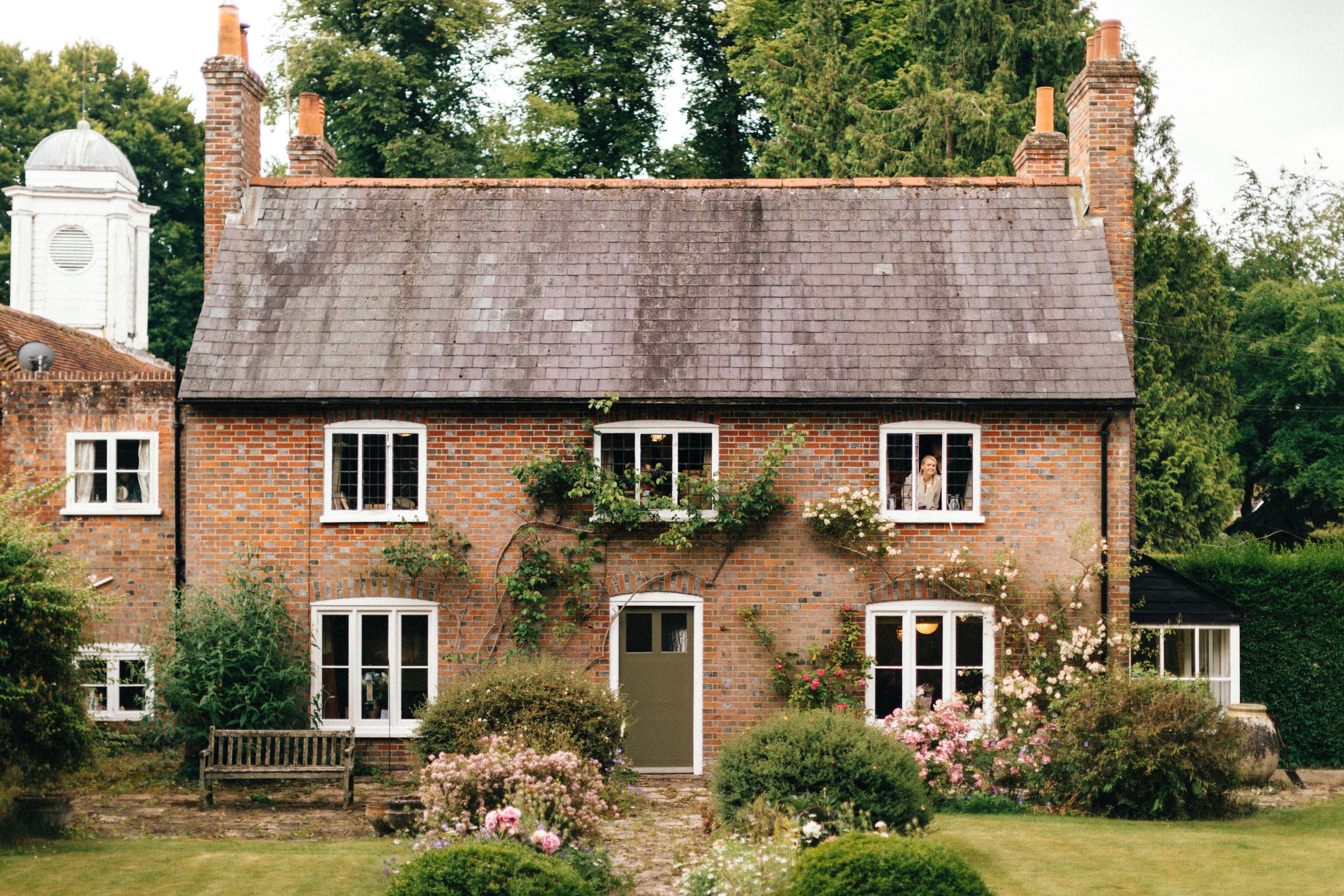 A two-story brick house with white-framed windows and a green front door, surrounded by a garden with blooming flowers and bushes, and trees in the background. A Bride smiles from the window.