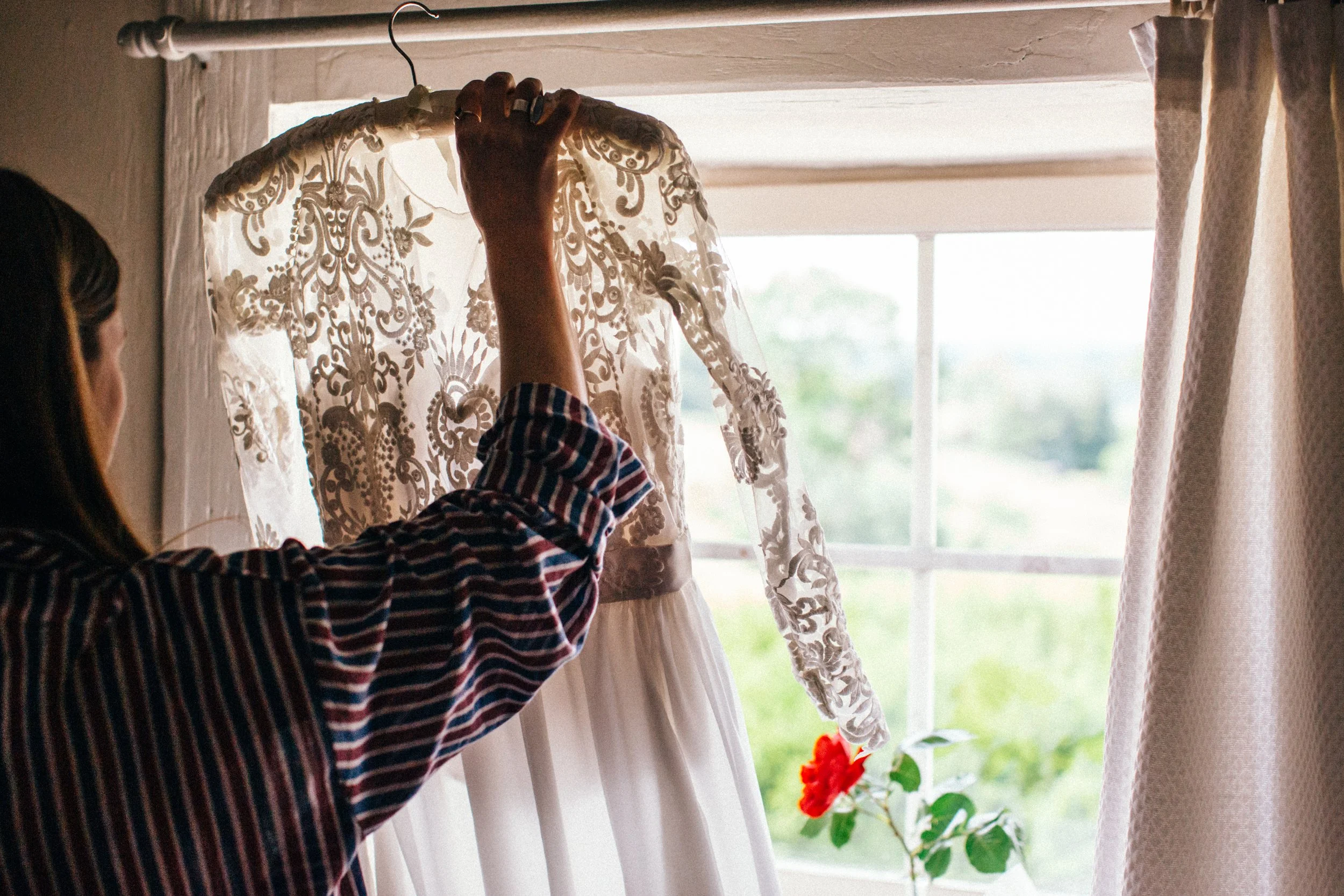 A woman with long dark hair, wearing a striped shirt, is holding a lace wedding dress on a hanger in front of a window with curtains, with sunlight coming through.