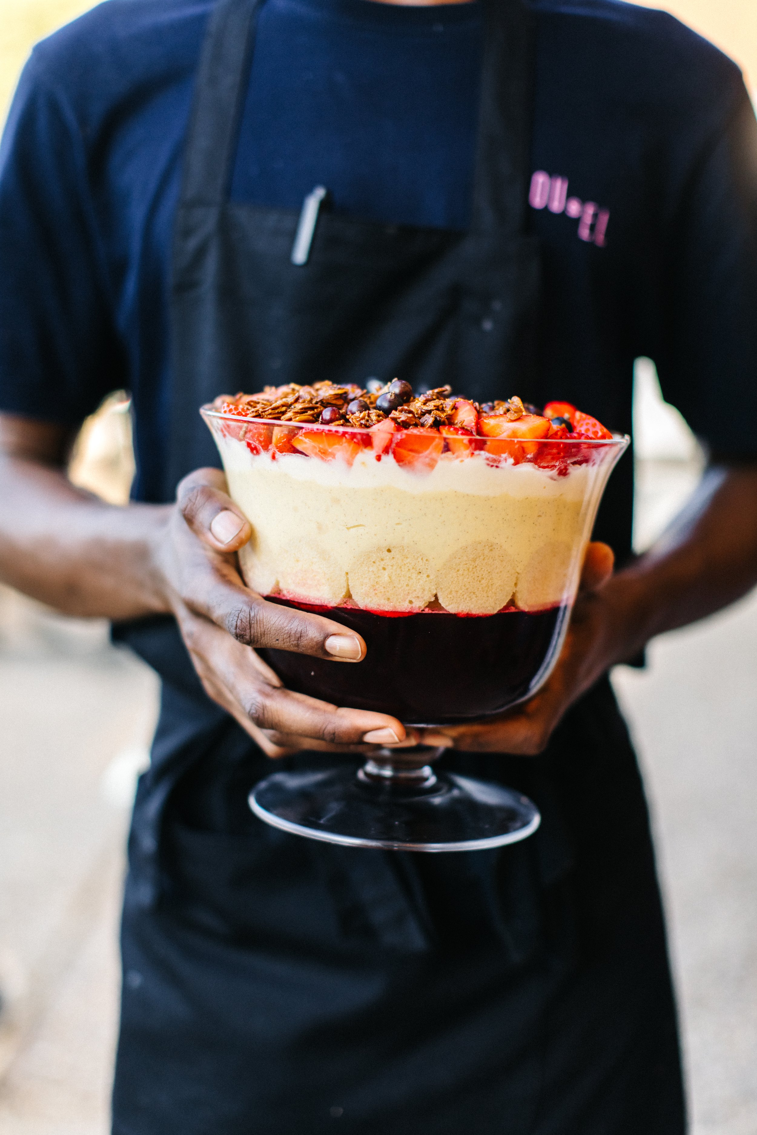 Person holding a glass bowl with a layered trifle made by Carousel topped with strawberries, chocolate, and other fruit.
