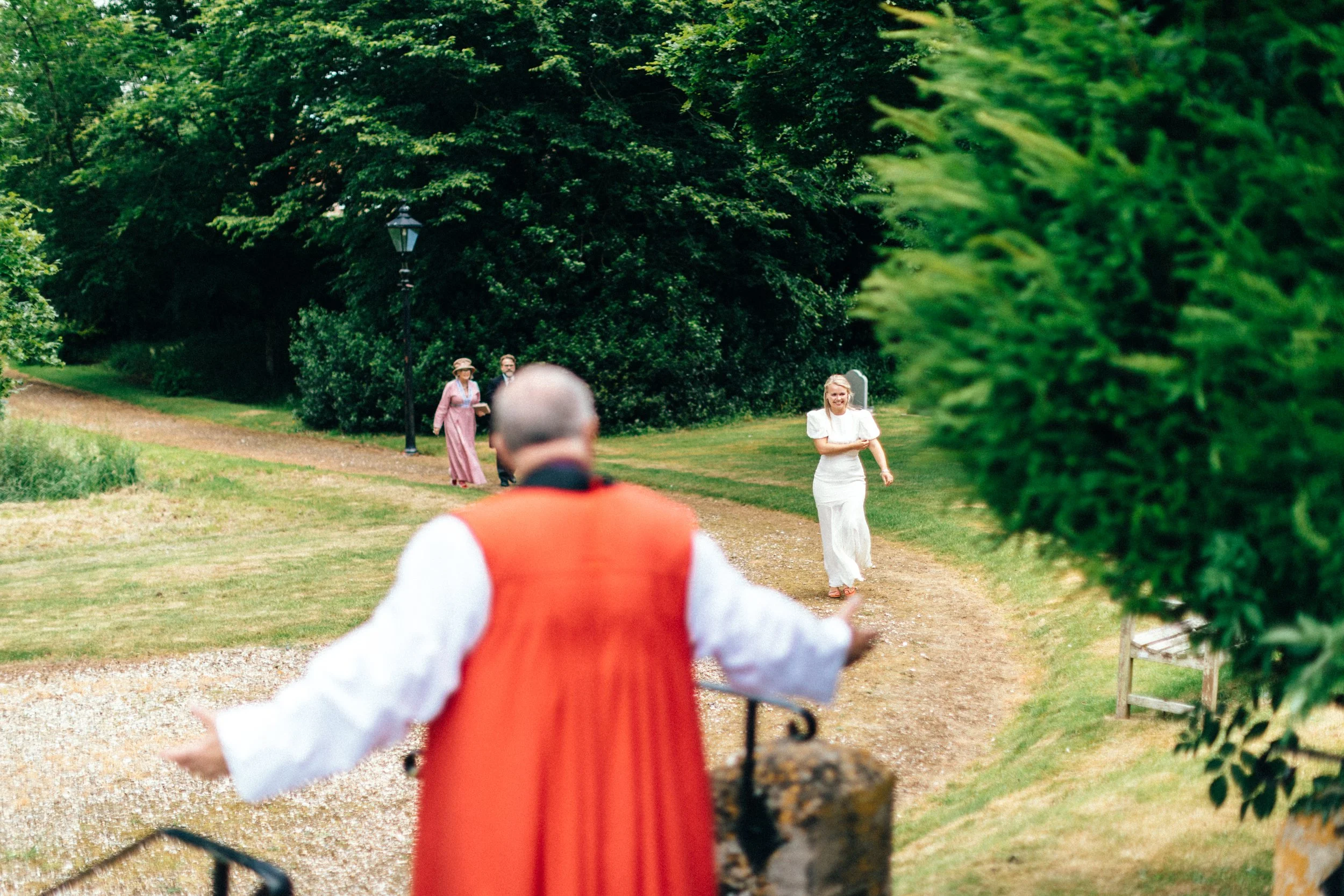 A bride in a white dress approaching a church laughing, with three people in vintage clothing in the background and a vicar in orange and white in the foreground.