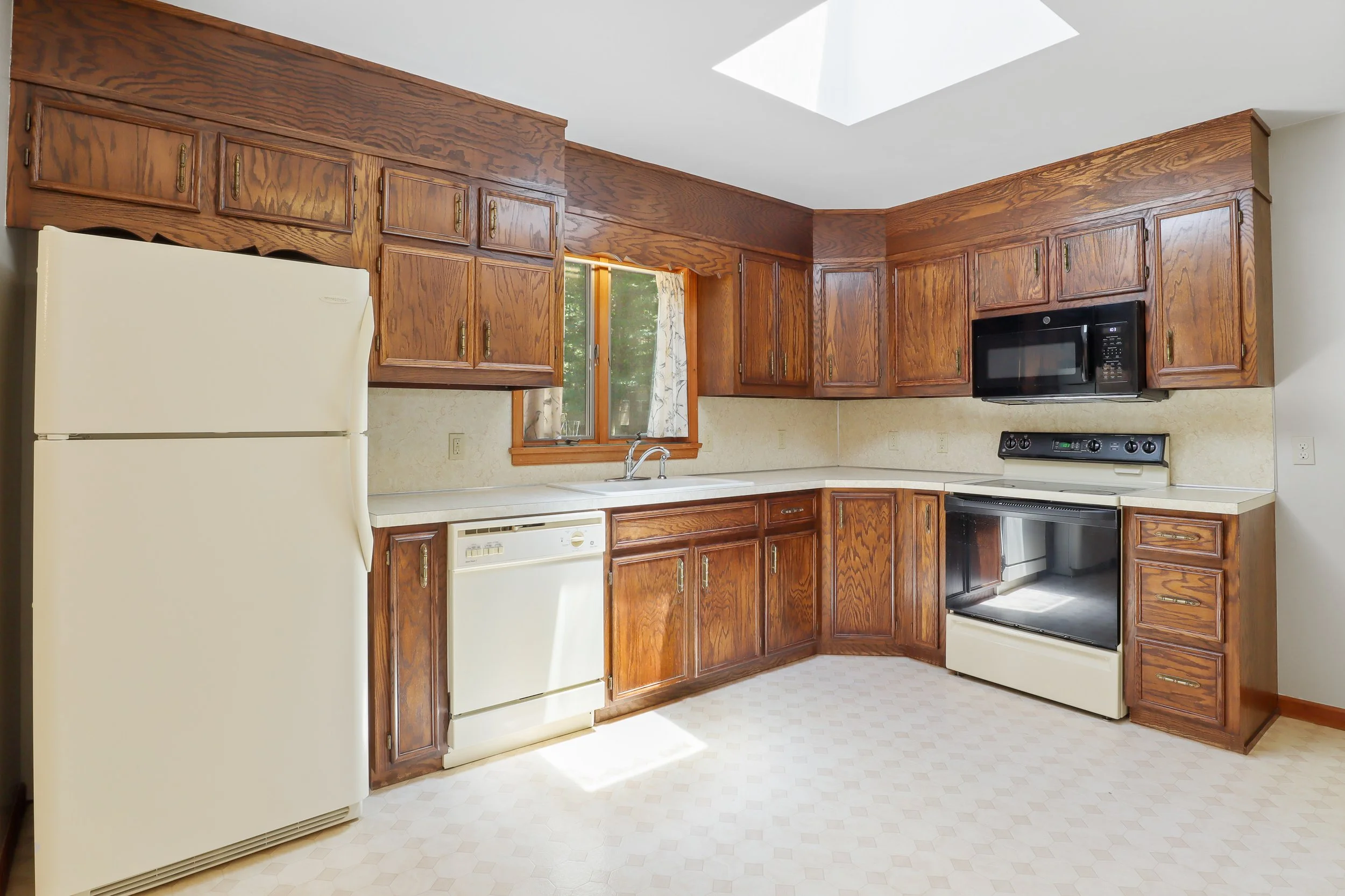 Kitchen with wooden cabinets, white appliances, window above sink, and skylight on ceiling.