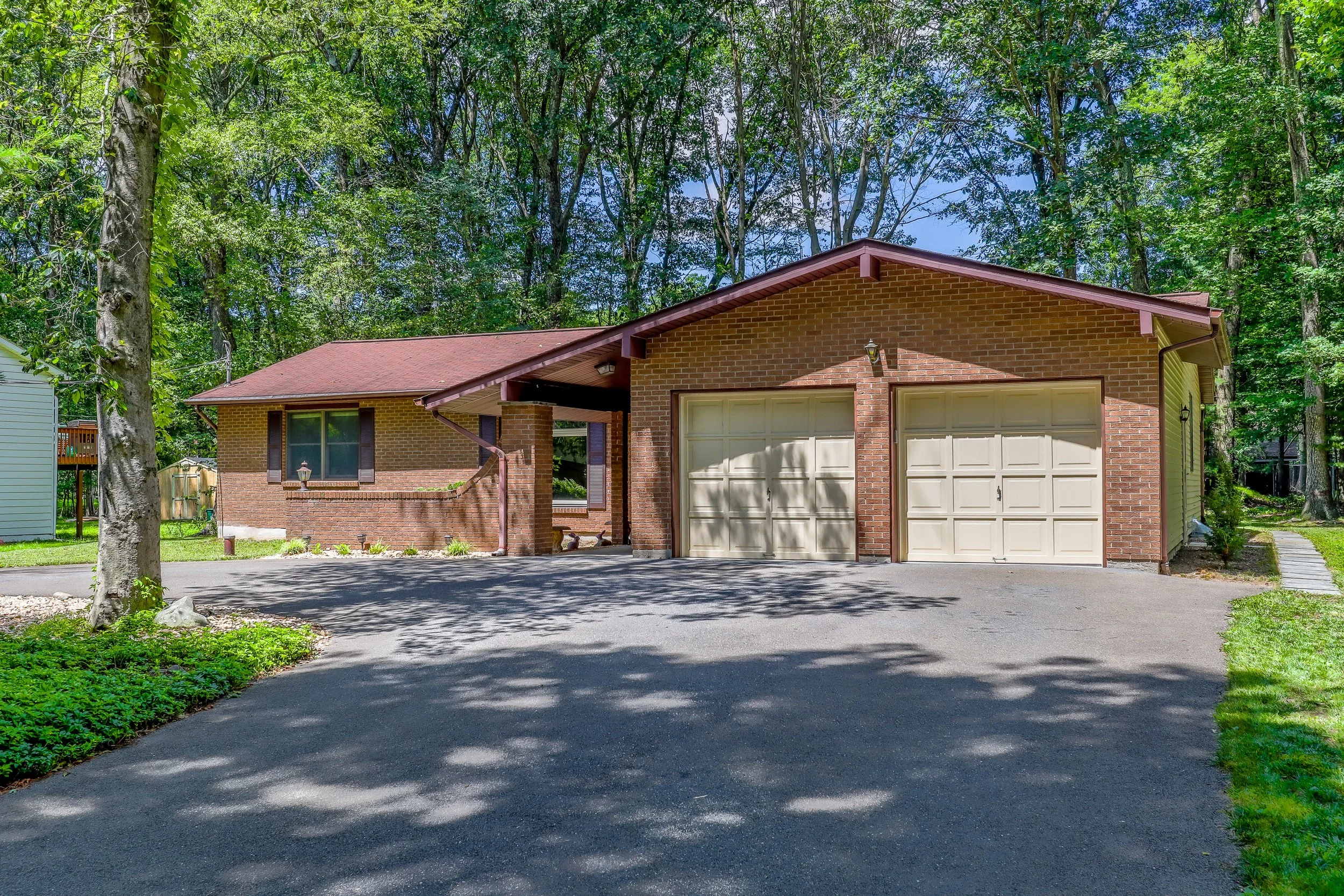A single-story brick house with a two-car garage, surrounded by trees and a paved driveway.