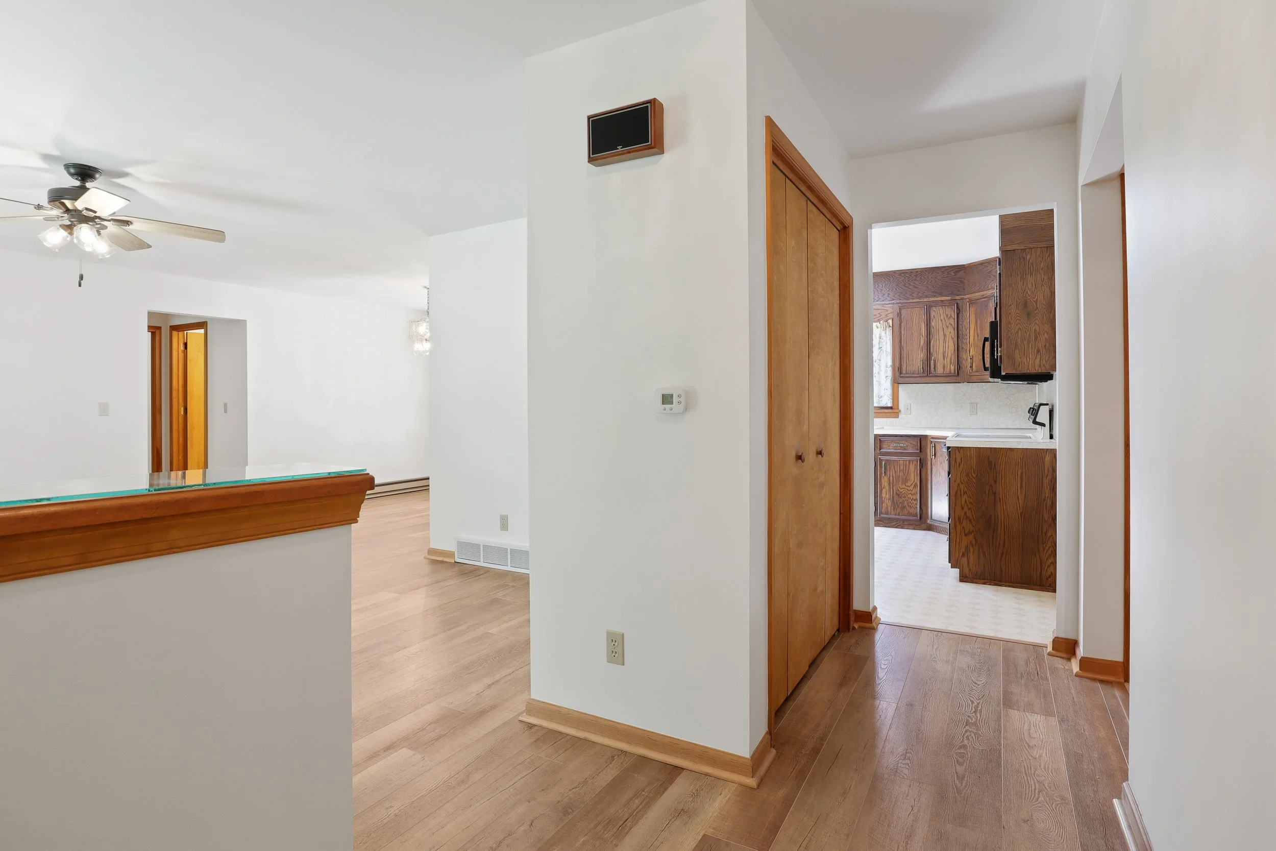 Interior view of a house showing a hallway with wooden flooring, a closet with wooden doors, and a kitchen with brown cabinets and white countertops.