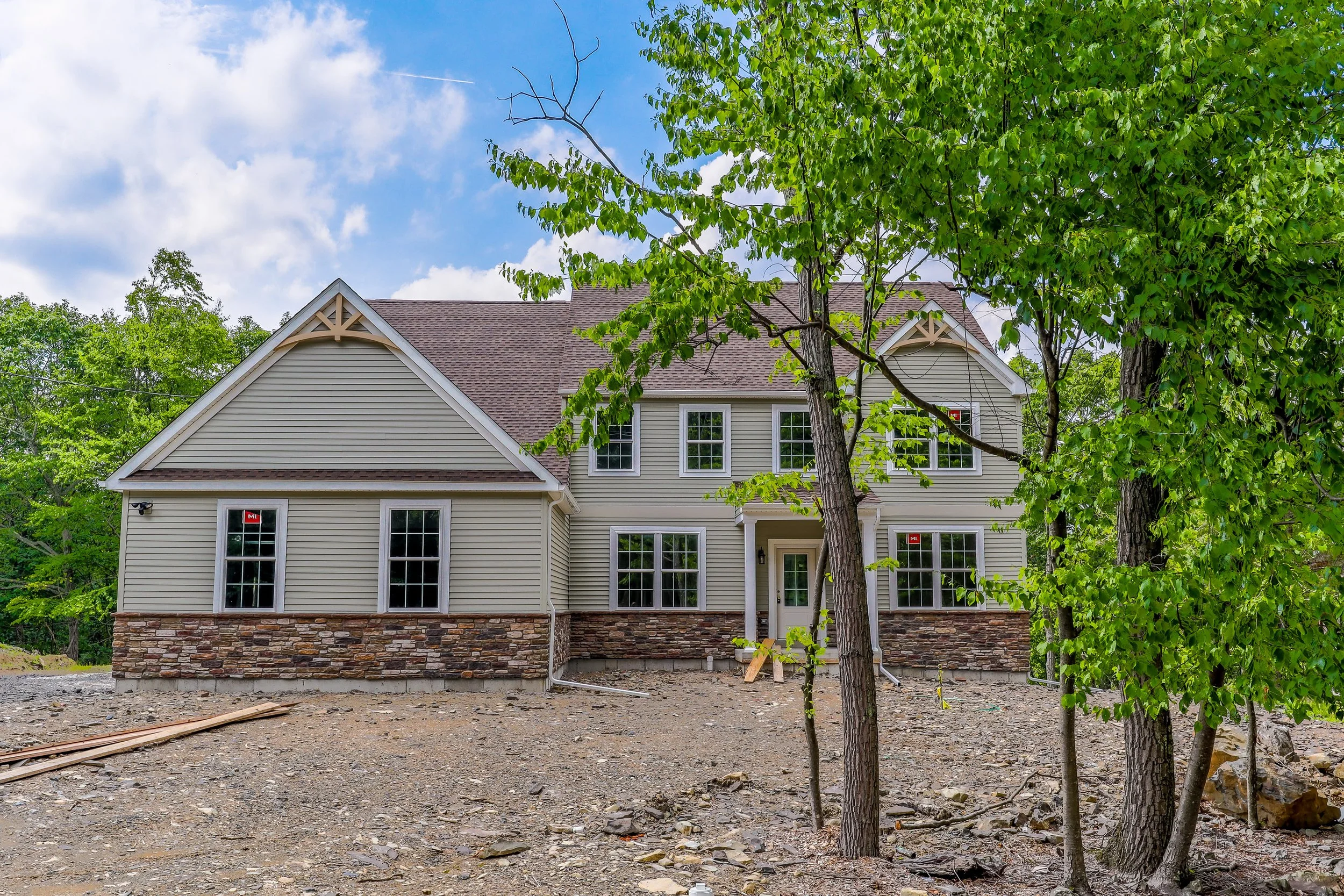 New two-story house under construction with vinyl siding, stone foundation, multiple windows, and a small front porch, surrounded by trees and a dirt yard under a partly cloudy sky.