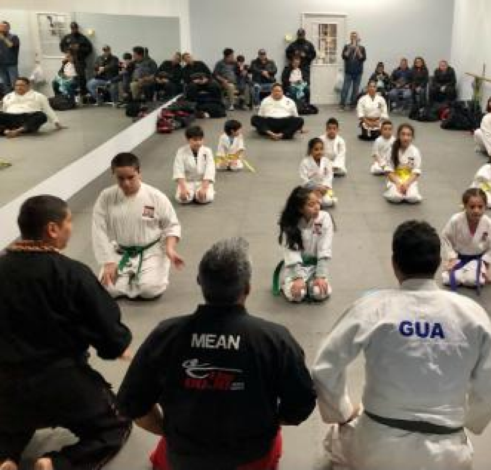 Karate students seated in rows facing instructors in a dojo, with an audience watching from the sides.