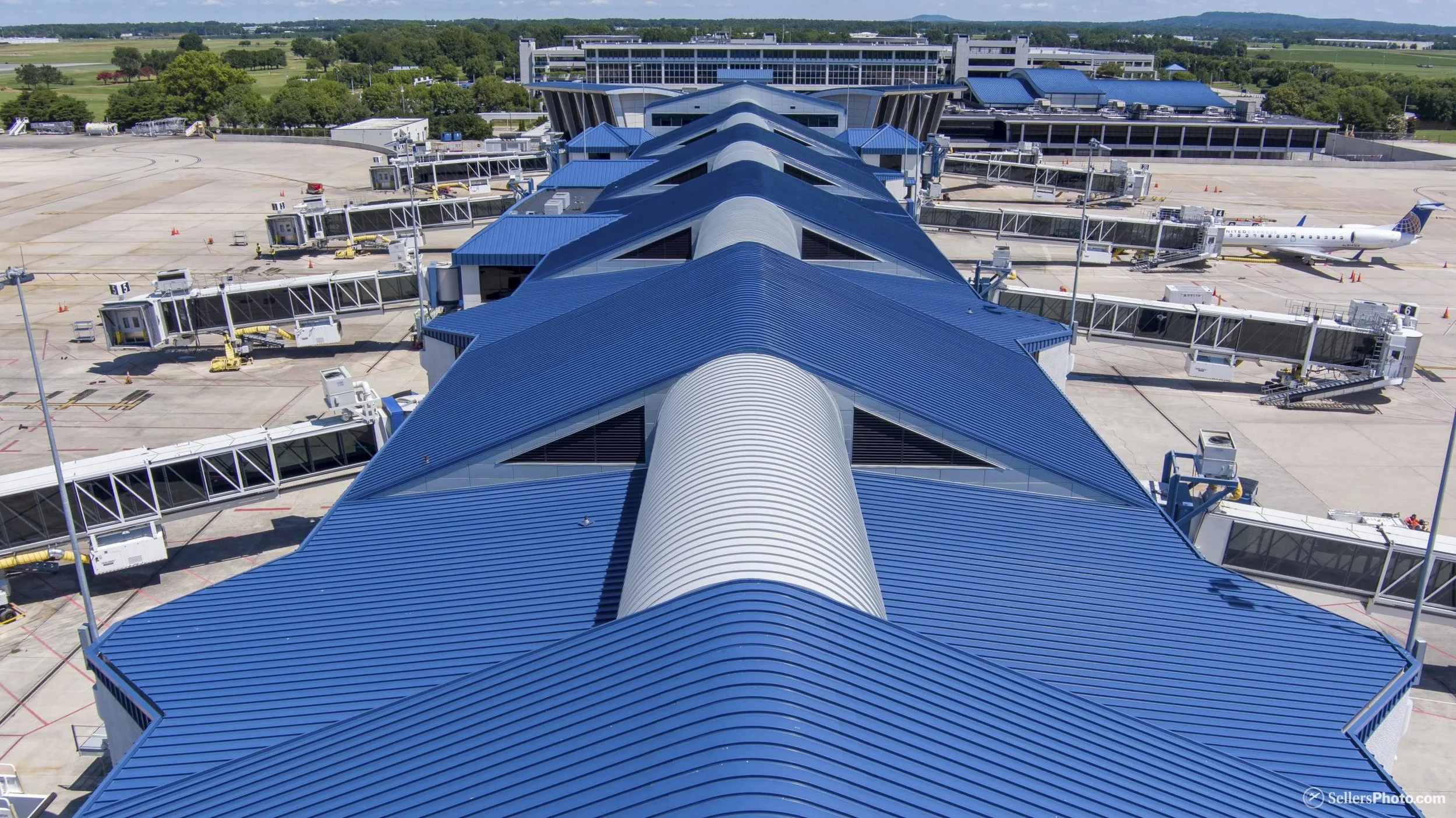 View of an airport terminal with a blue metal roof and connected jet bridges, with an airplane parked at the gate in the background, surrounded by tarmac and open sky.