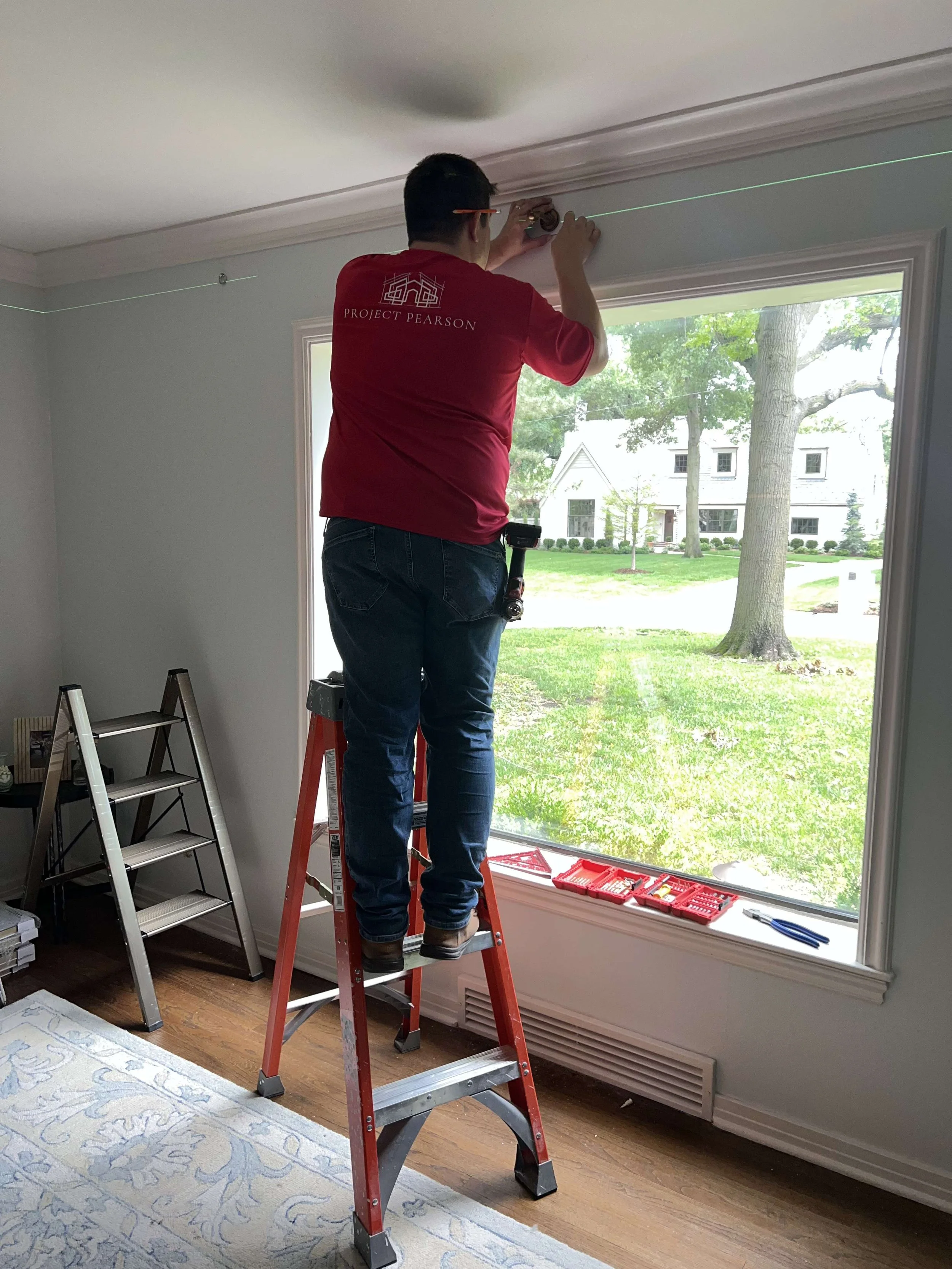 A person standing on a ladder installing or repairing a laser level on the ceiling next to a large window in a room with hardwood floors and a patterned rug.