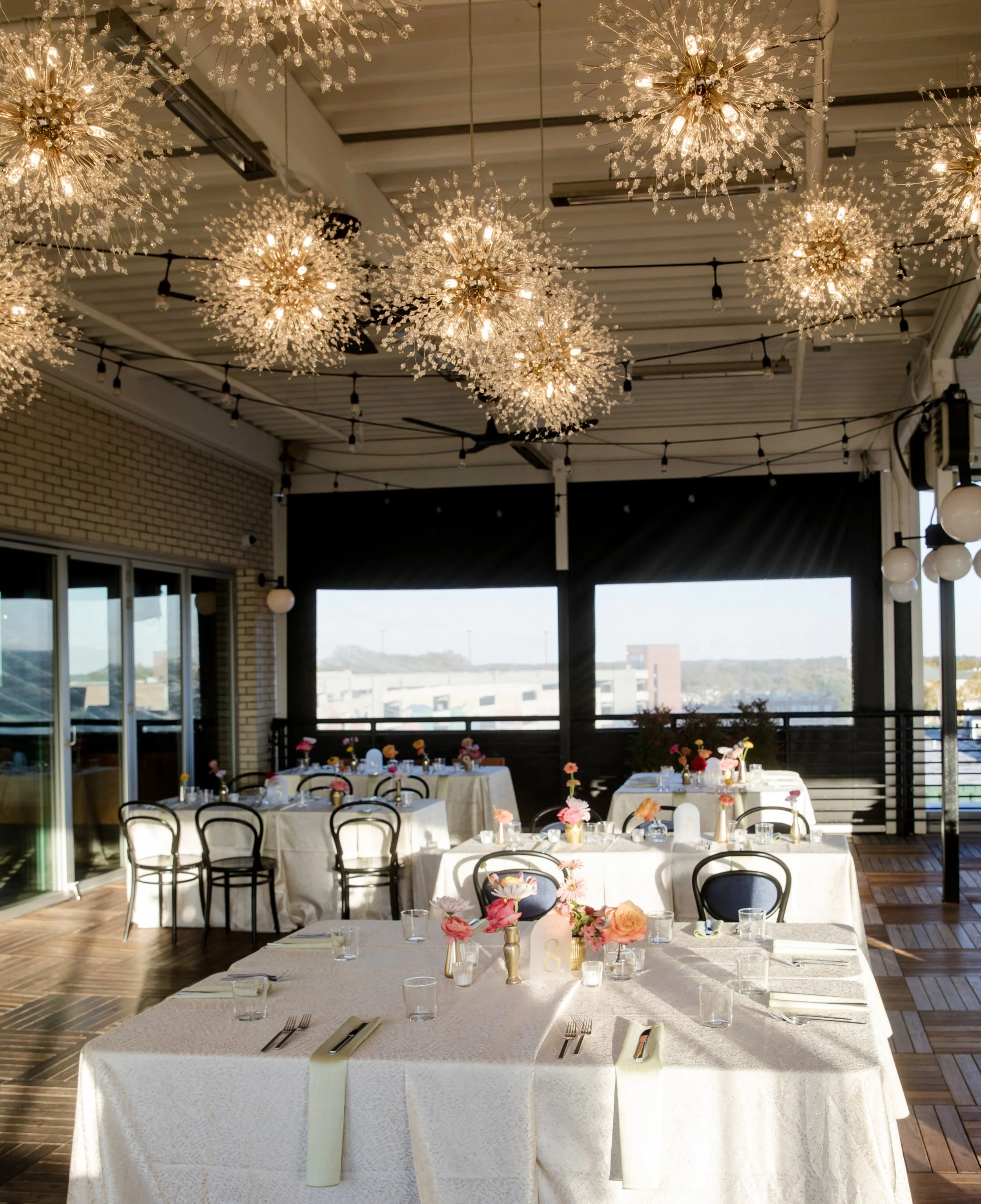 Indoor event space decorated for a celebration with white tables, floral centerpieces, and ornate chandeliers hanging from the ceiling.
