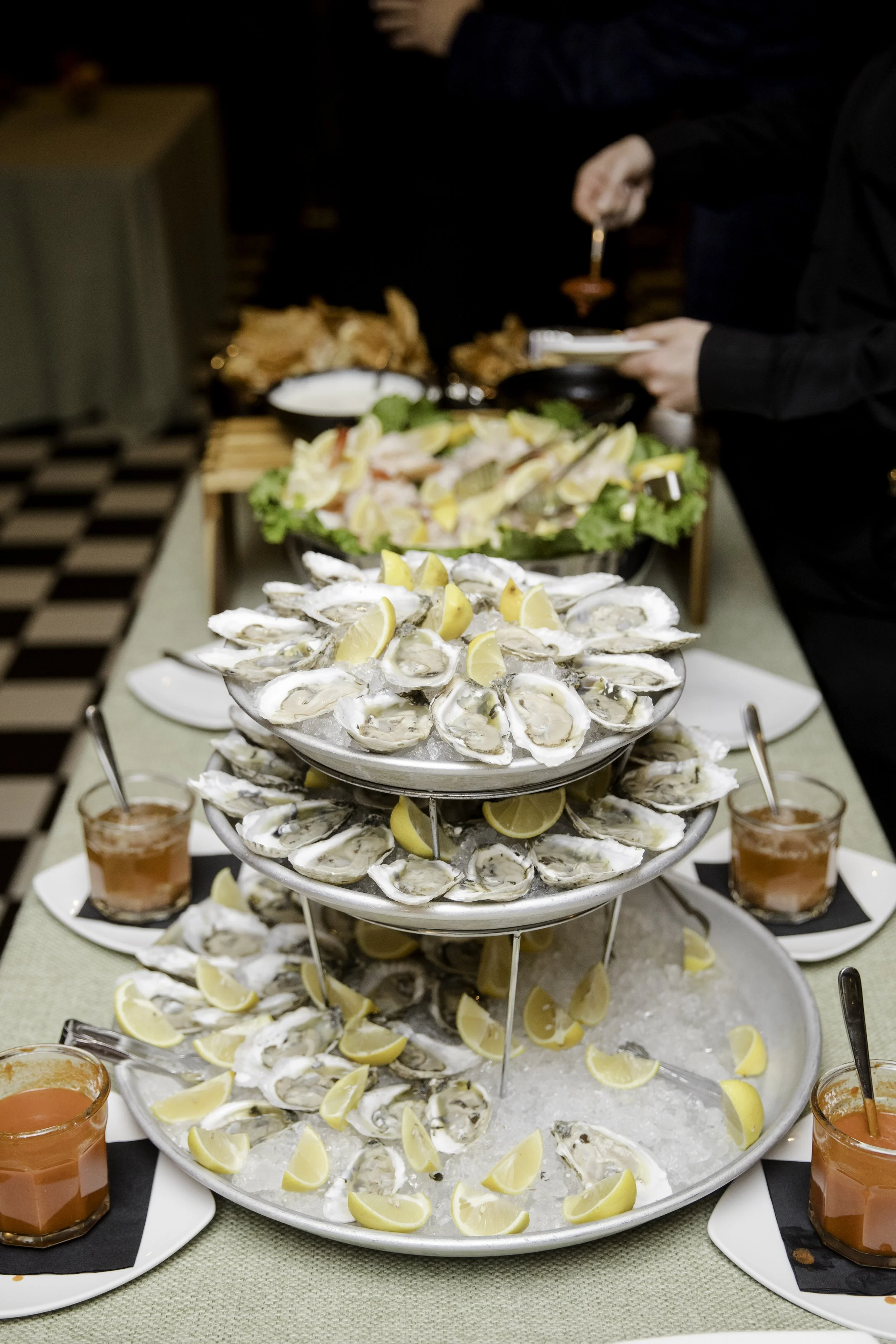 A tiered display of oysters with lemon wedges, with small glasses of tomato-based drink on the side, on a banquet table at a seafood buffet.