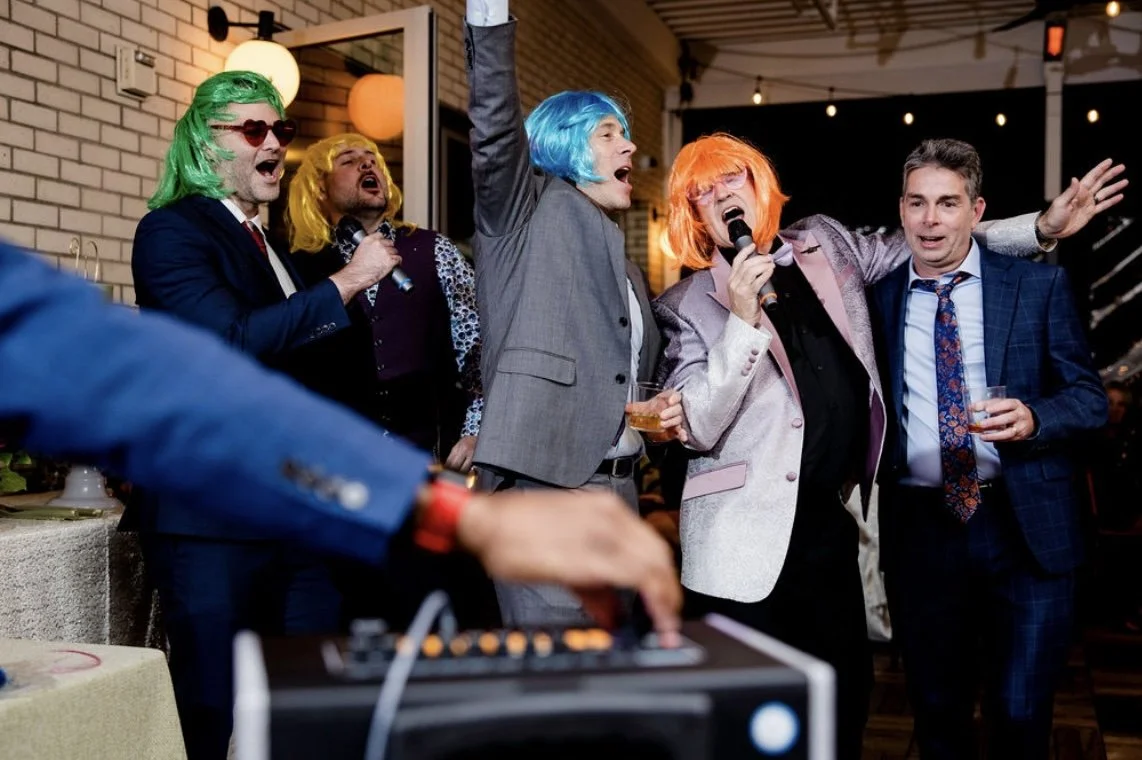 Group of people at a party wearing colorful wigs, singing karaoke, and enjoying drinks, with a DJ in the foreground.