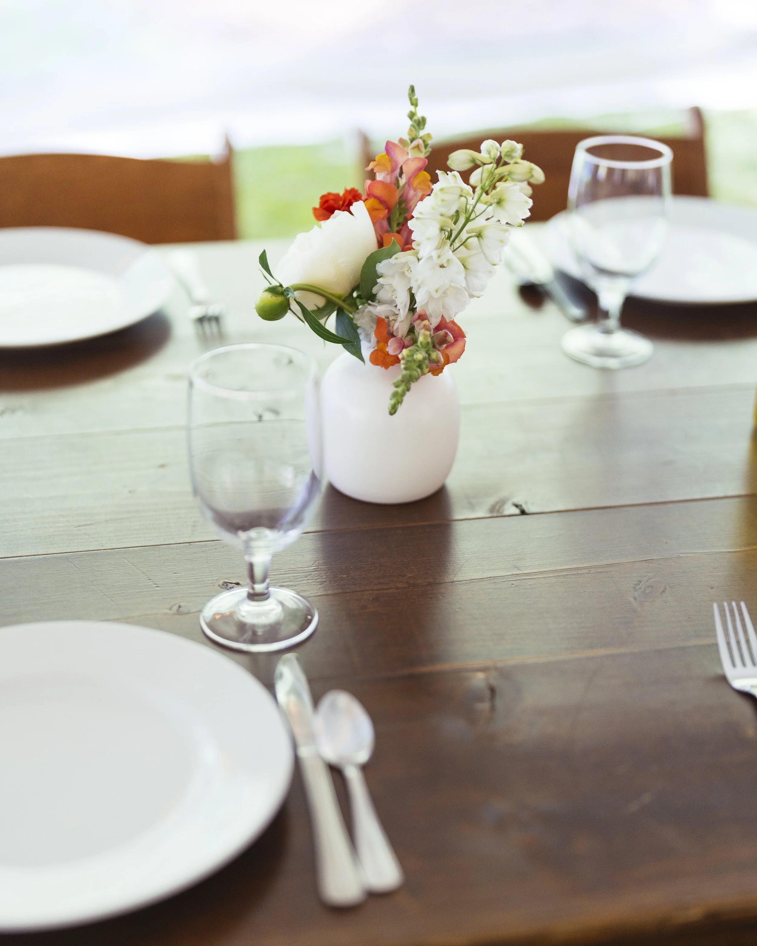 A dining table set for a meal with a white vase holding a flower arrangement of white, pink, orange, and green flowers; a wine glass, water glass, white plate, spoon, and fork are visible.