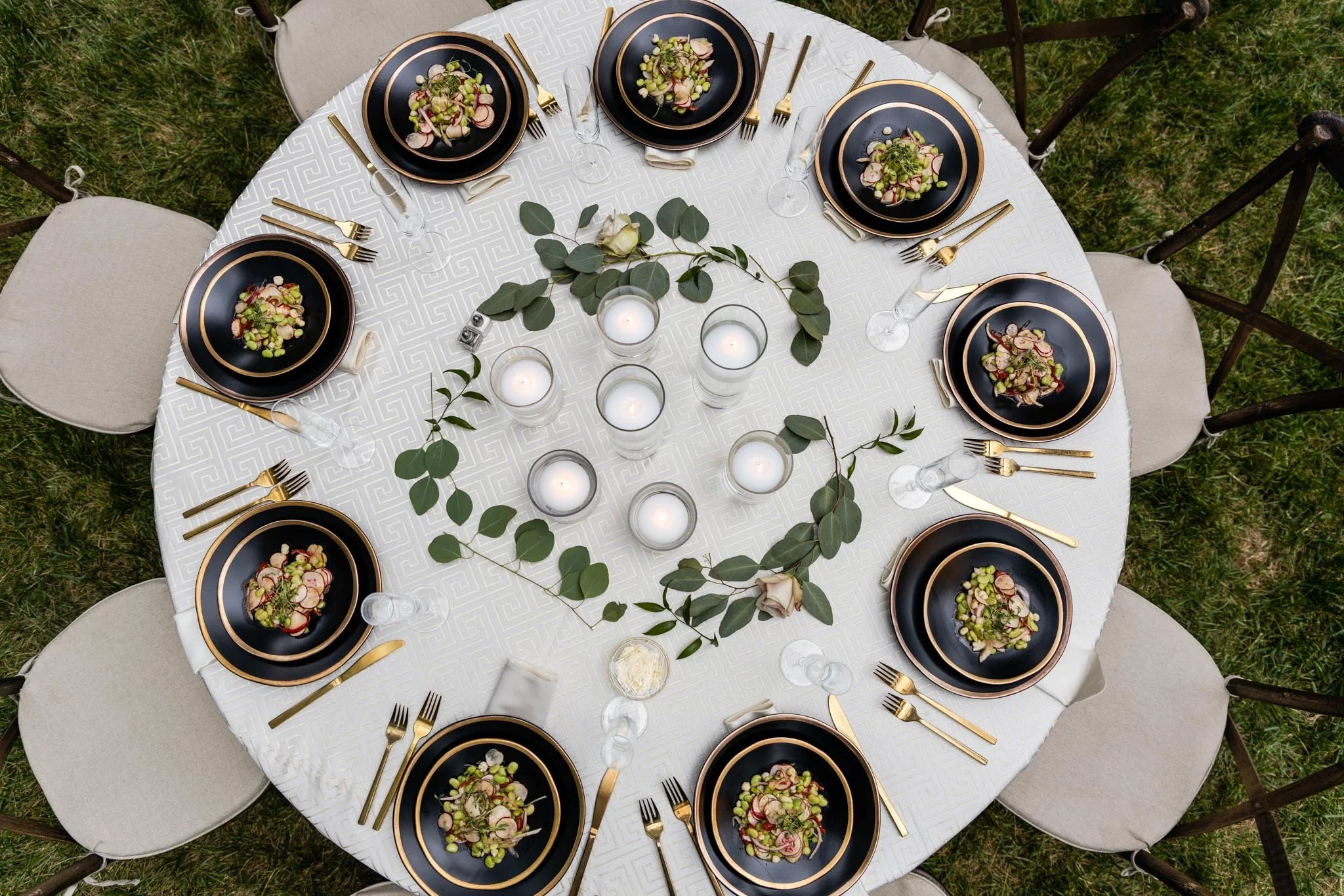 An elegant outdoor table setting with black and gold dinnerware, salad dishes, gold flatware, wine glasses, and white candles surrounded by green leaves on a white tablecloth, set on grass.