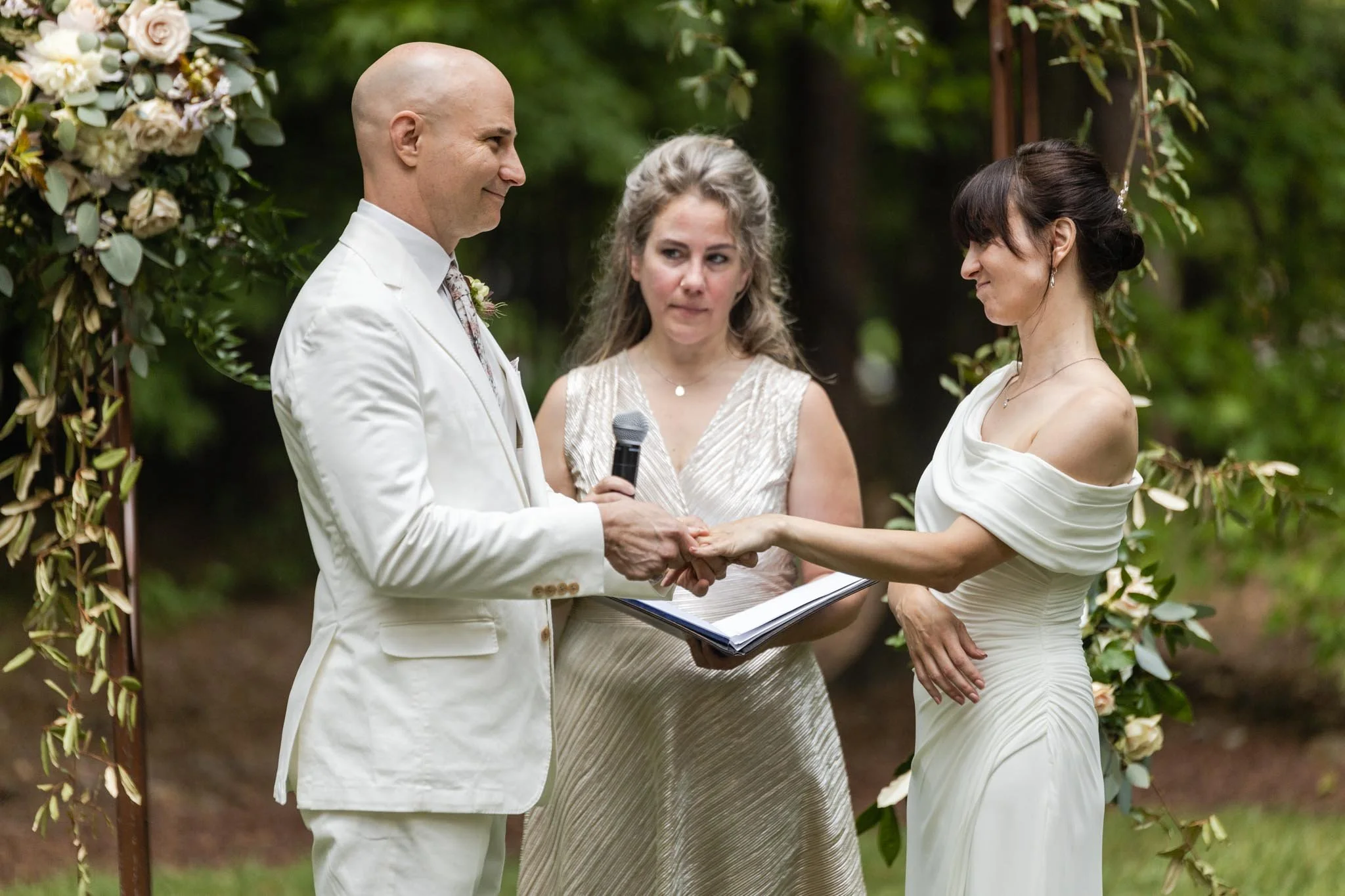 A couple getting married outdoors, holding hands and gazing at each other during their wedding ceremony, with an officiant standing behind them, in front of an arch decorated with flowers.