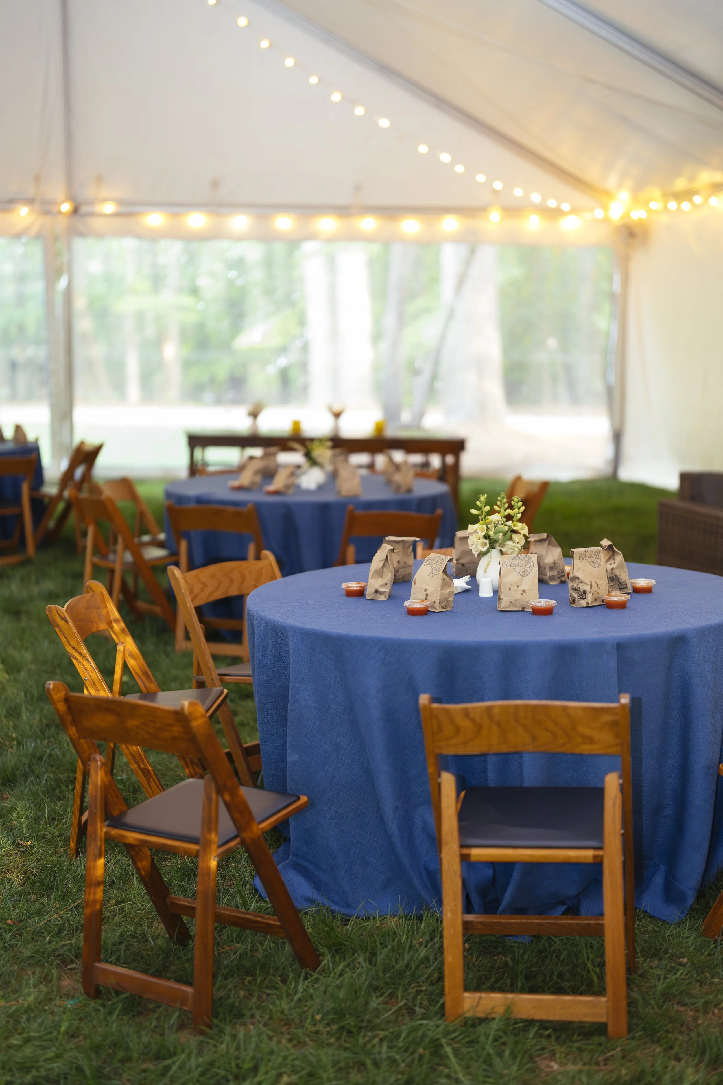 Outdoor event setup inside a large white tent with string lights, round tables covered with blue tablecloths, and wooden chairs on green grass, decorated with small flower arrangements and paper bags with food.