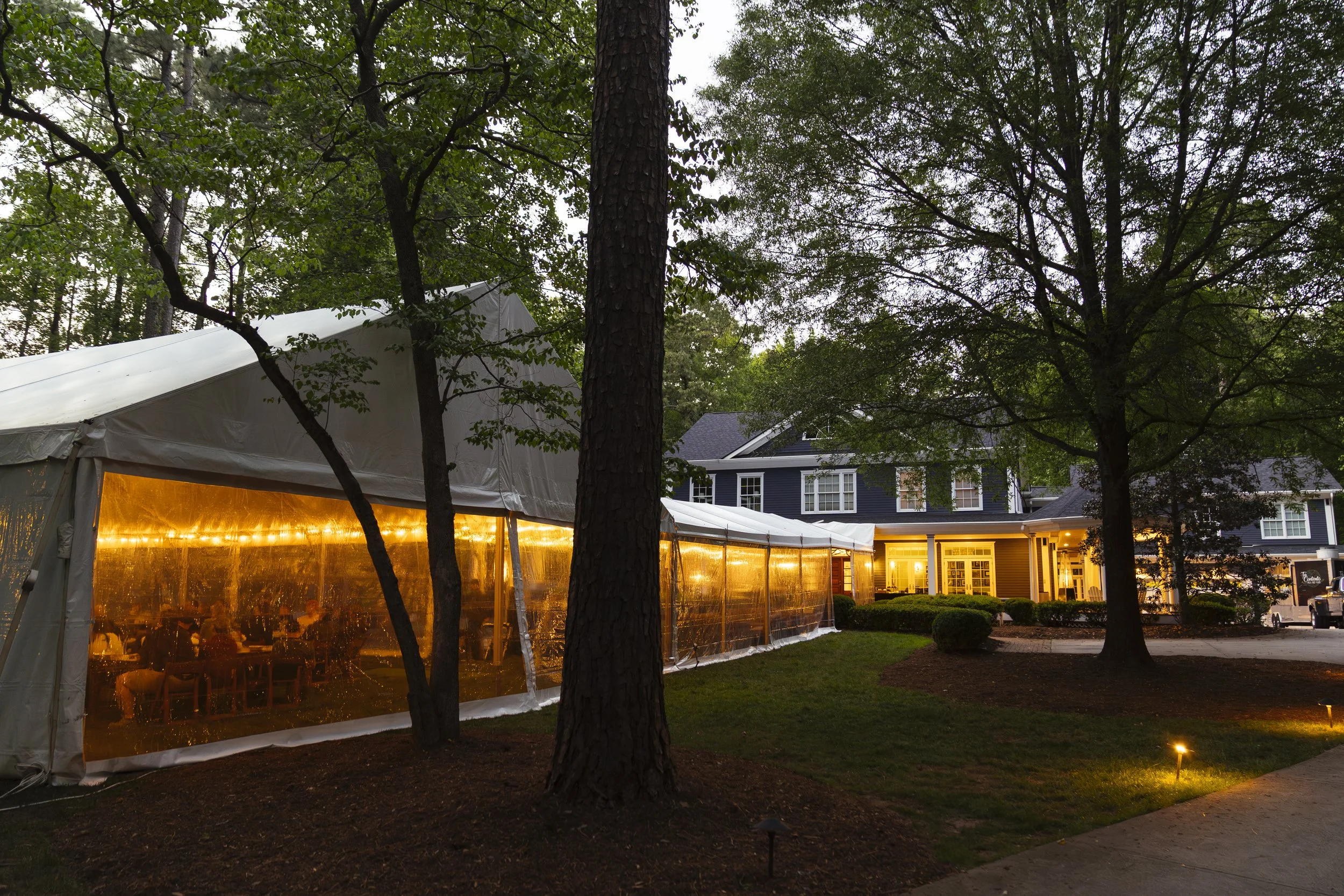 An outdoor wedding reception area with a large white tent illuminated with warm yellow lights, surrounded by trees and residential buildings at dusk.