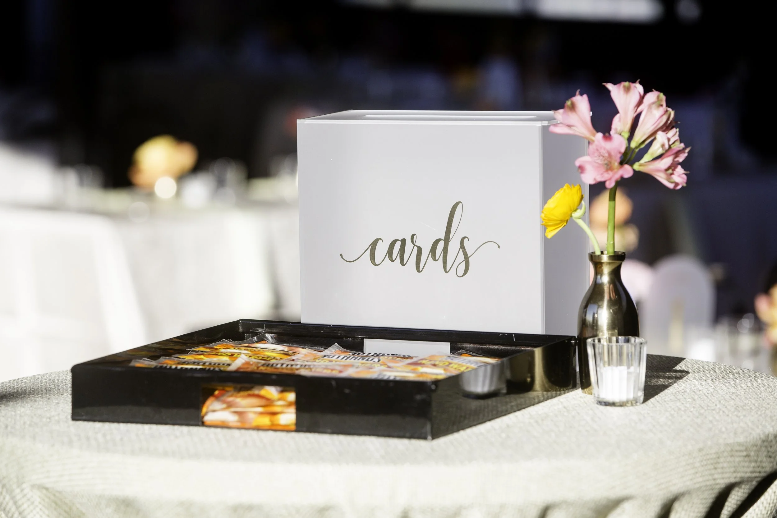 A white box with the word 'cards' written on it in cursive is placed on a black tray. To the right, there is a small vase with pink and yellow flowers. The scene appears to be part of a decorative table setting, possibly at an event or celebration.