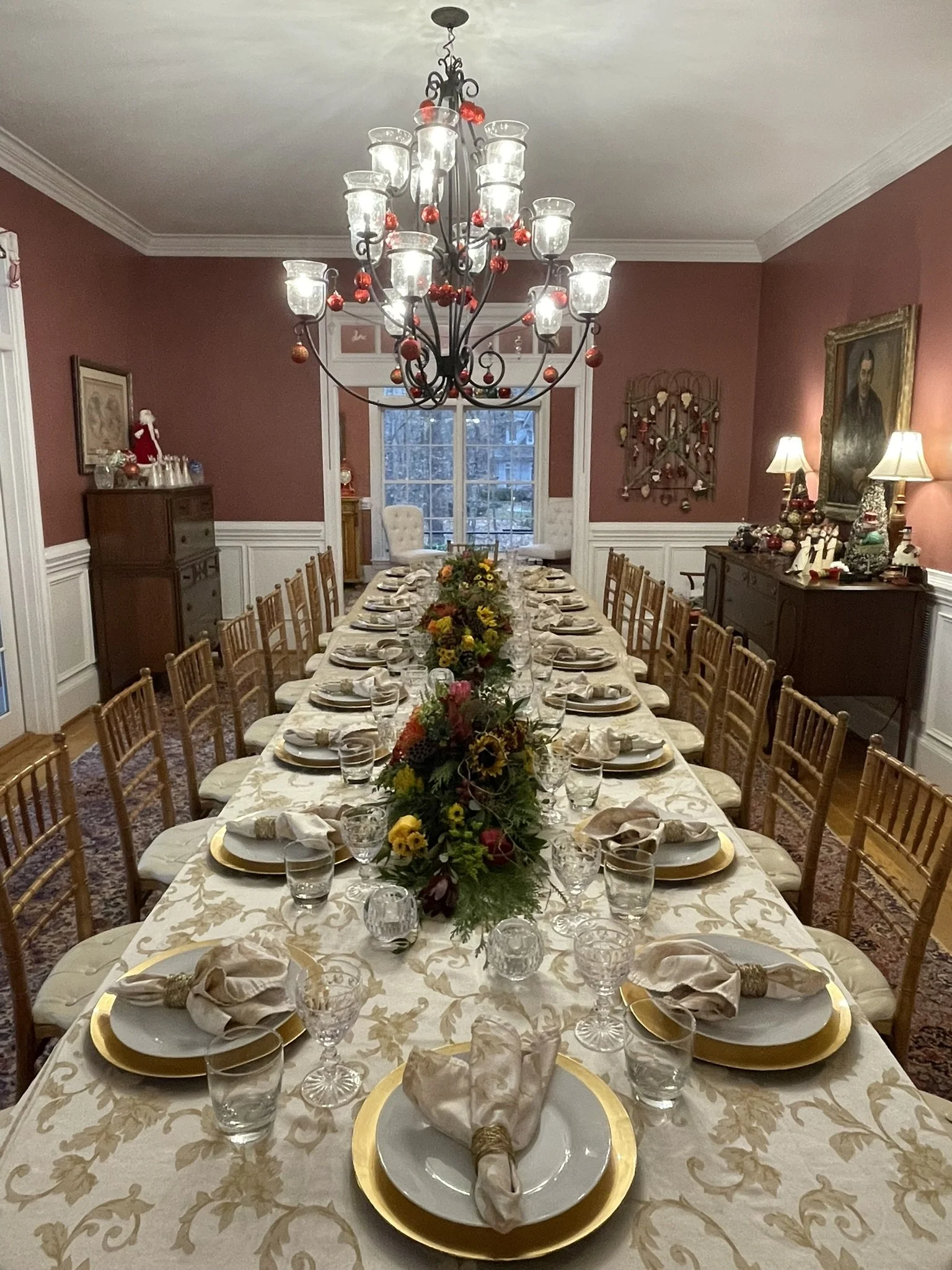 A large dining room with a long table set for a meal, decorated with a centerpiece of flowers and surrounded by gold-rimmed plates, glasses, and napkins. There is a chandelier hanging from the ceiling and warm lighting from a side cabinet and wall la