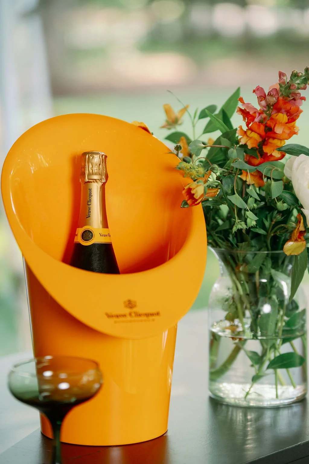 A bottle of Veuve Clicquot champagne inside a yellow ice bucket, next to a vase of colorful flowers on a table.
