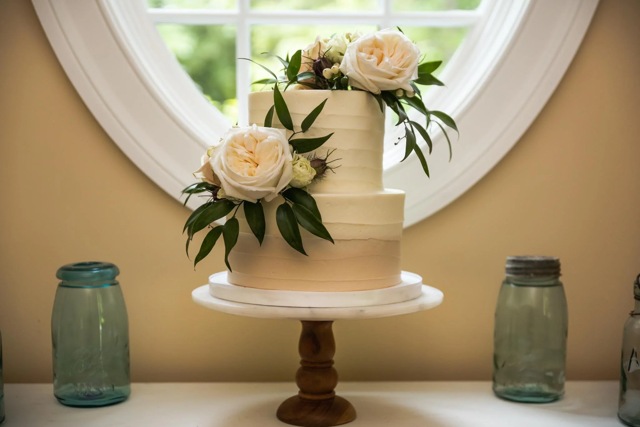A white wedding cake decorated with white roses, greenery, and small white berries, placed on a wooden stand in front of a window.