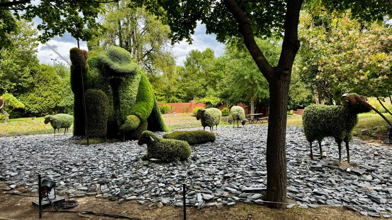 Topiary sculptures of a dog with a stick, smaller dogs, and sheep in a park setting, surrounded by trees and greenery with a gravel ground cover.