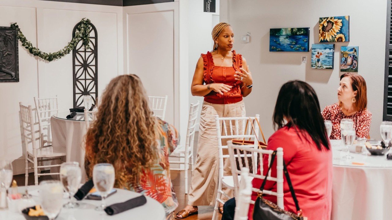 A woman standing and speaking to a seated group at a restaurant or event space, with paintings on the wall and decorated with a garland.