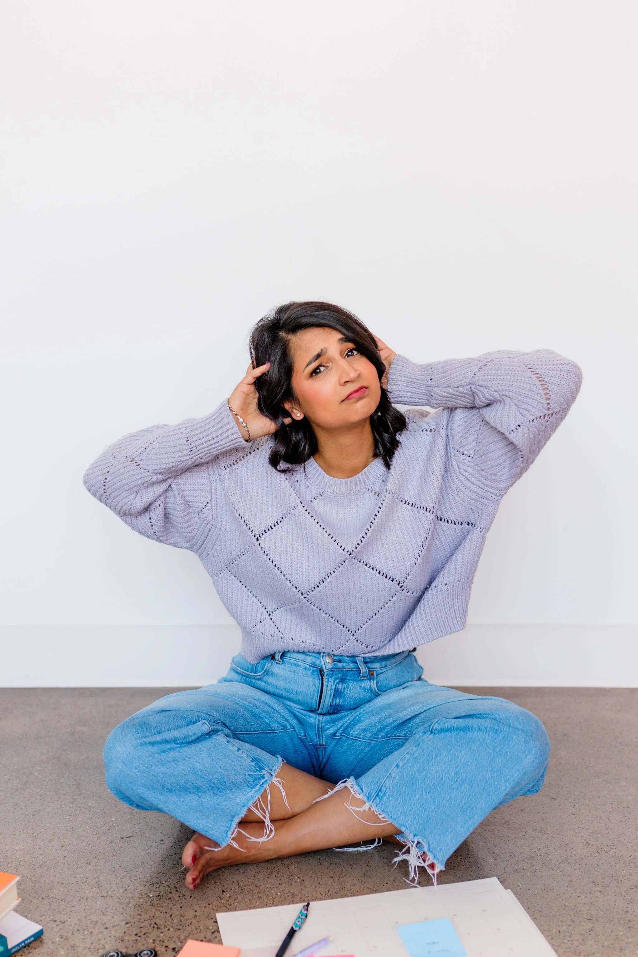 Woman sitting cross-legged on the floor, holding her head and looking confused, with papers spread around.